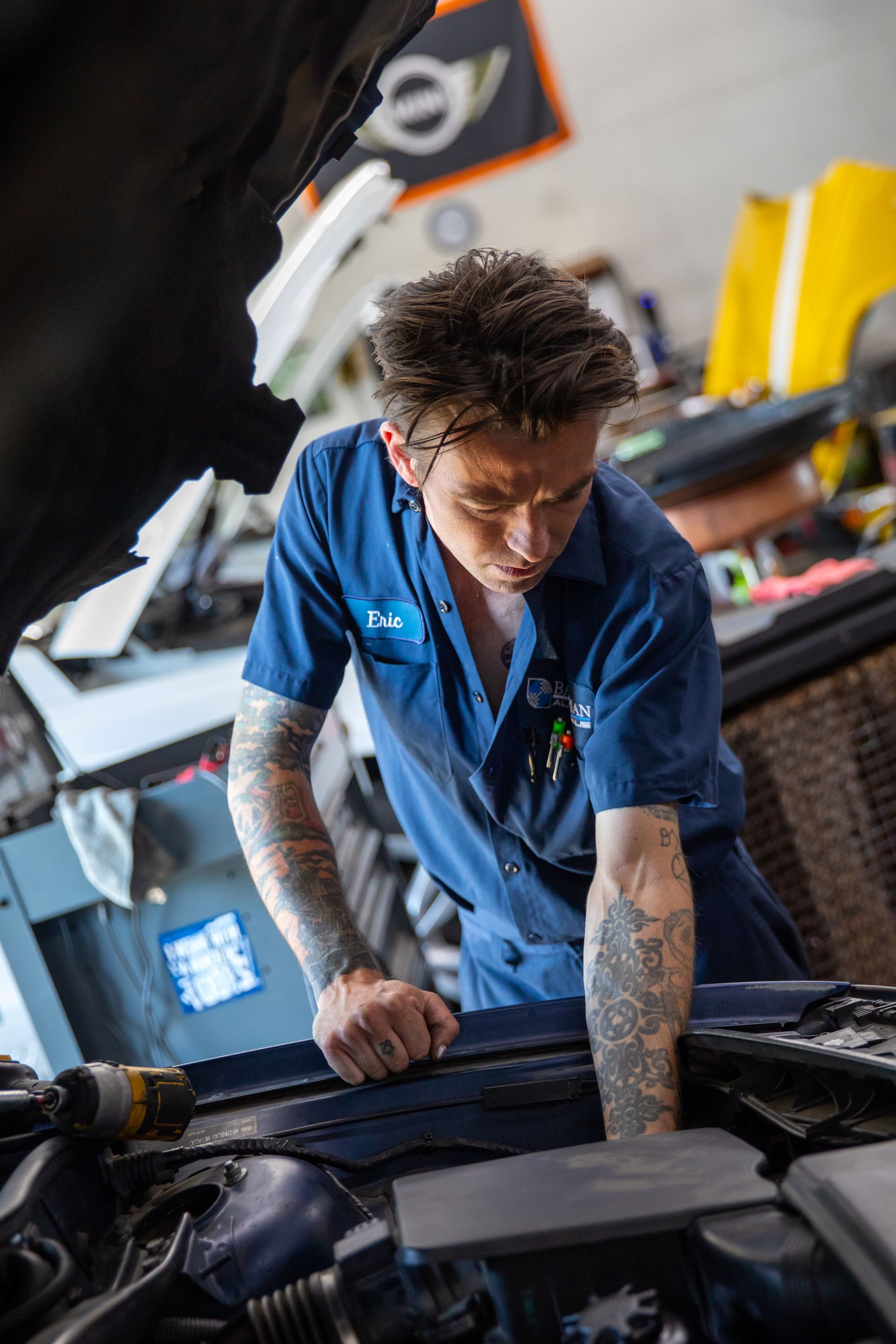 A tattooed mechanic in a blue uniform leans over an open car engine, focused on working under the hood in a garage. | Bavarian Auto Haus