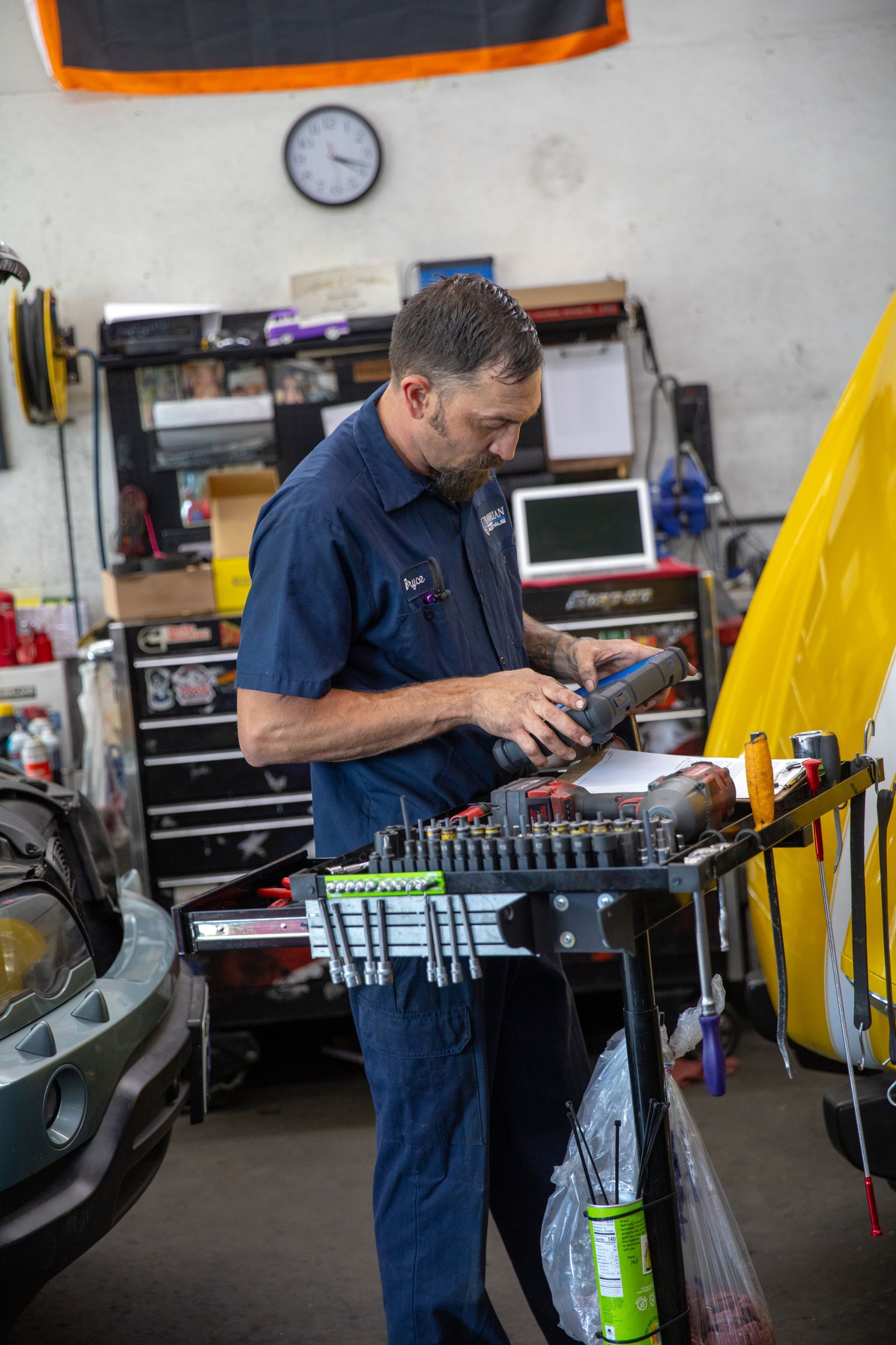 A technician in a blue uniform reviews a diagnostic scanner while standing at a rolling tool cart in an auto shop. | Bavarian Auto Haus