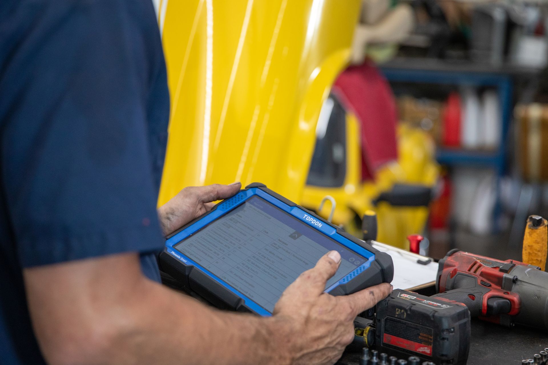 Mechanic holding a tablet to diagnose a vehicle in a garage with a yellow car hood visible in the background. | Bavarian Auto Haus