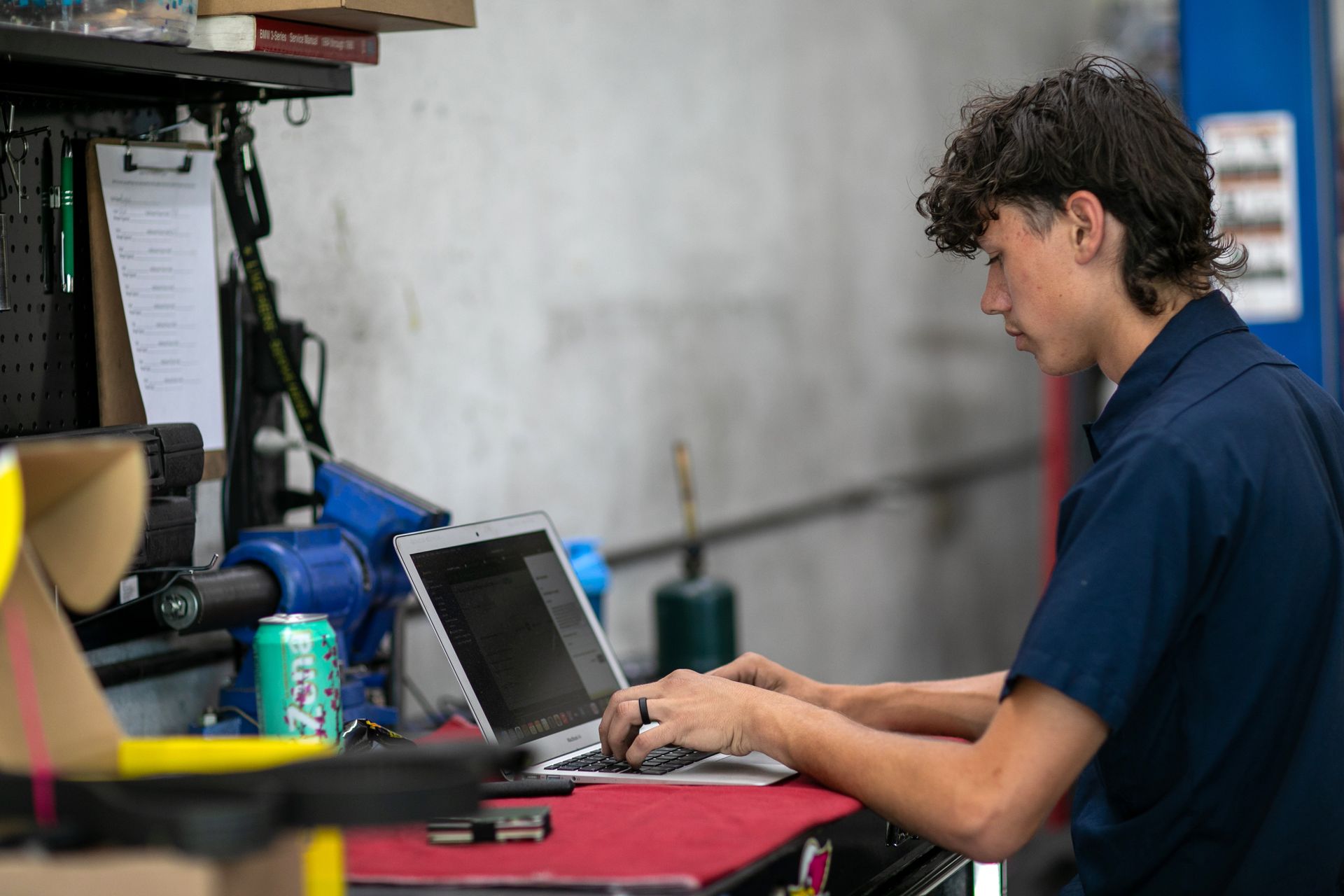 A person in a blue shirt works on a laptop at a workbench in a cluttered workshop. | Bavarian Auto Haus