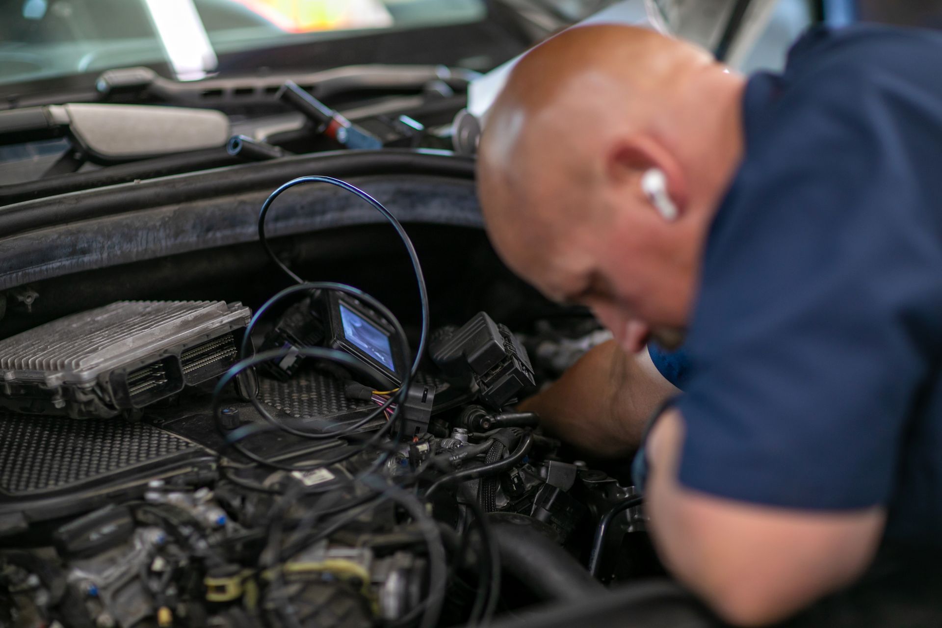 A mechanic in a blue shirt leans over an open car engine, using a handheld diagnostic tool to inspect the components. | Bavarian Auto Haus