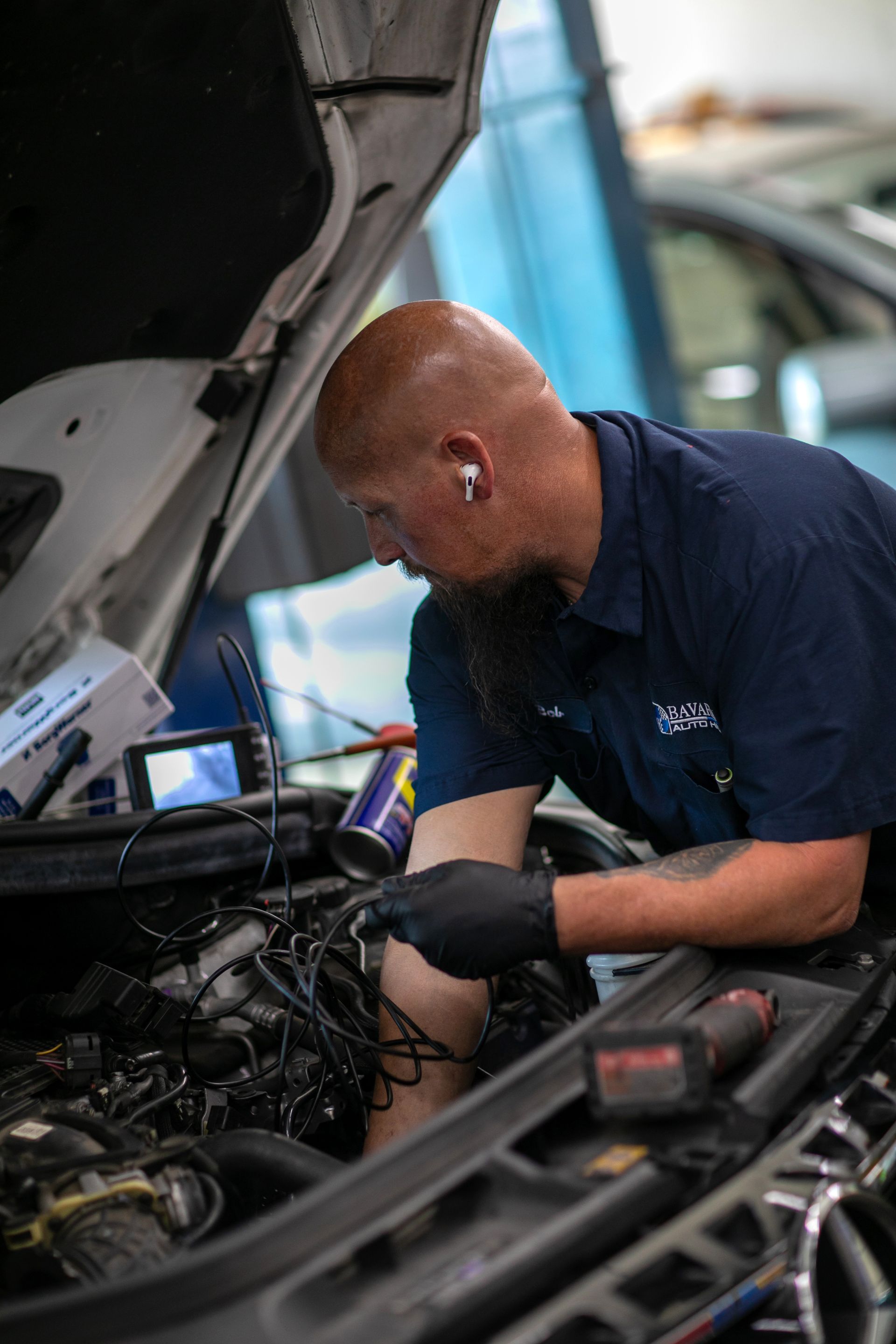 A technician in a blue uniform and black gloves works on the engine of a car with the hood raised in an auto shop. | Bavarian Auto Haus
