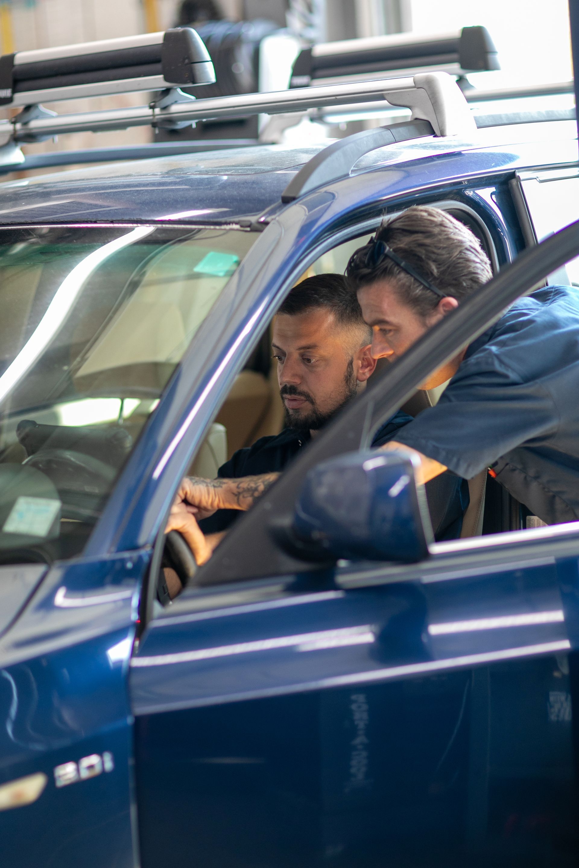 Two mechanics in blue uniforms look at the steering wheel of a blue car inside an auto repair shop. | Bavarian Auto Haus