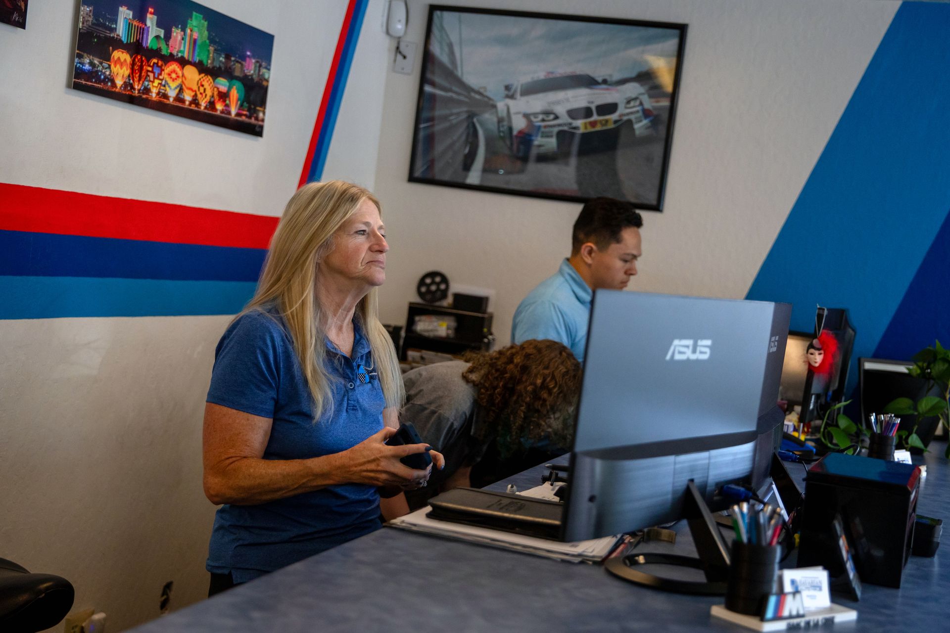 Two people work at desks in an office with blue and red wall accents and automotive wall art. | Bavarian Auto Haus