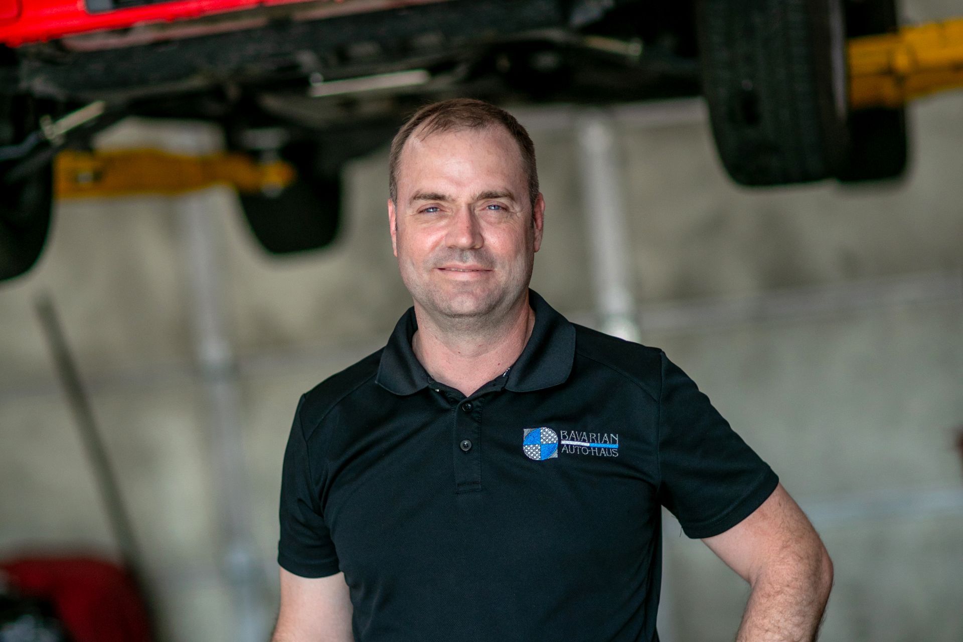 A person in a black polo shirt stands in an automotive workshop, with a vehicle hoisted on a lift in the background. | Bavarian Auto Haus
