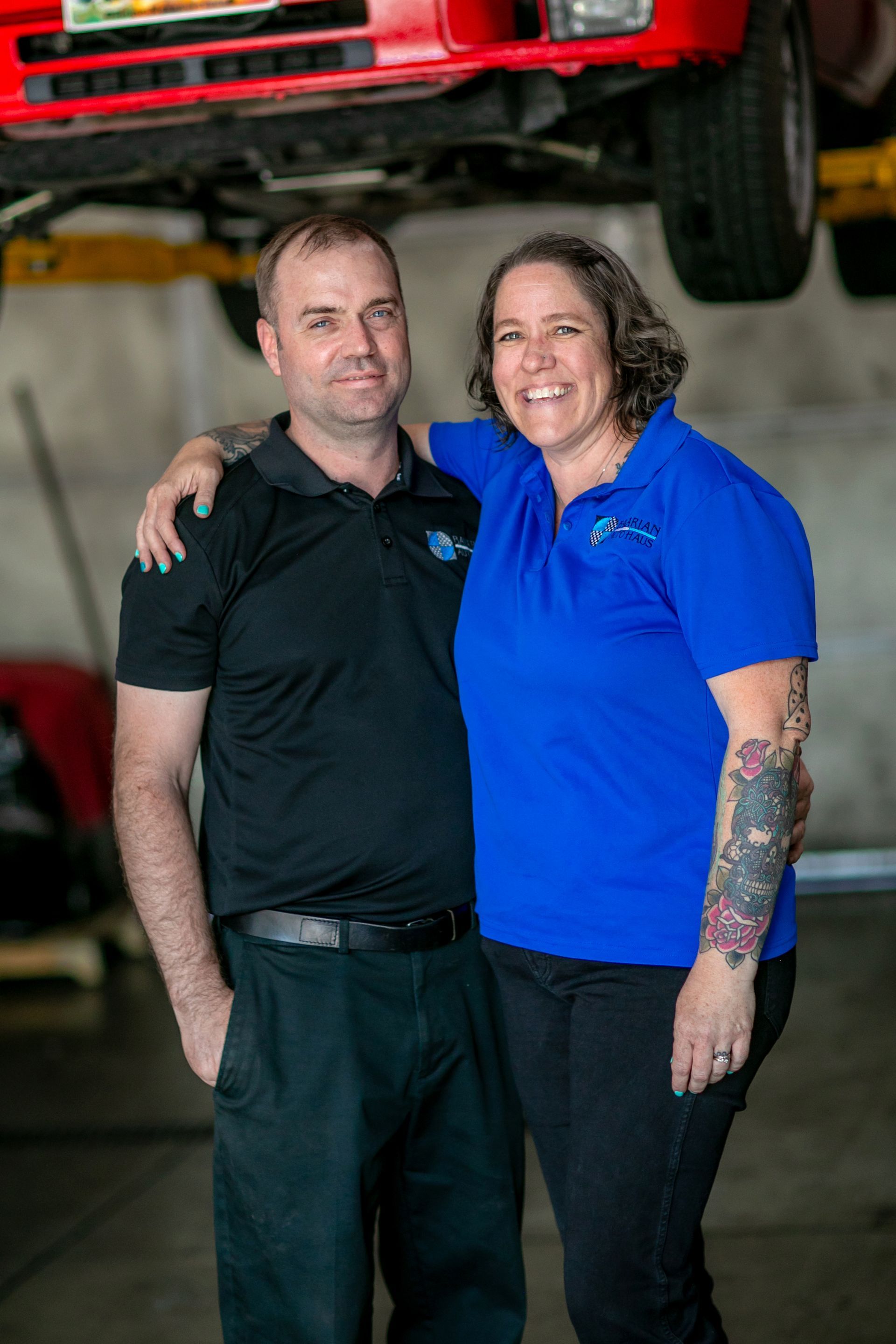 A smiling couple stands in an auto repair shop, posed in front of a red car lifted on a maintenance rack. | Bavarian Auto Haus