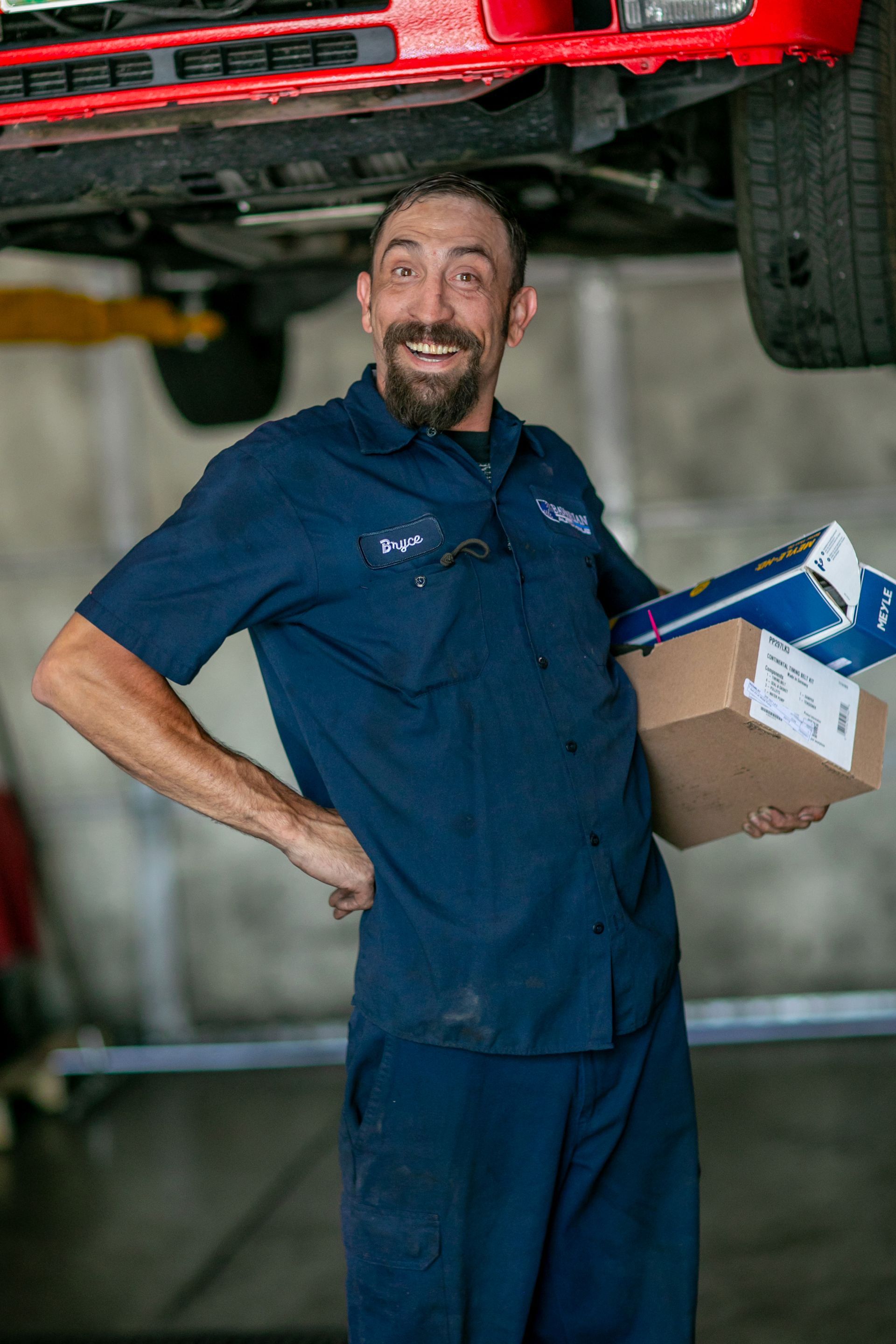 A smiling mechanic in a navy uniform holds auto parts boxes in an auto repair shop with a car lifted in the background. | Bavarian Auto Haus