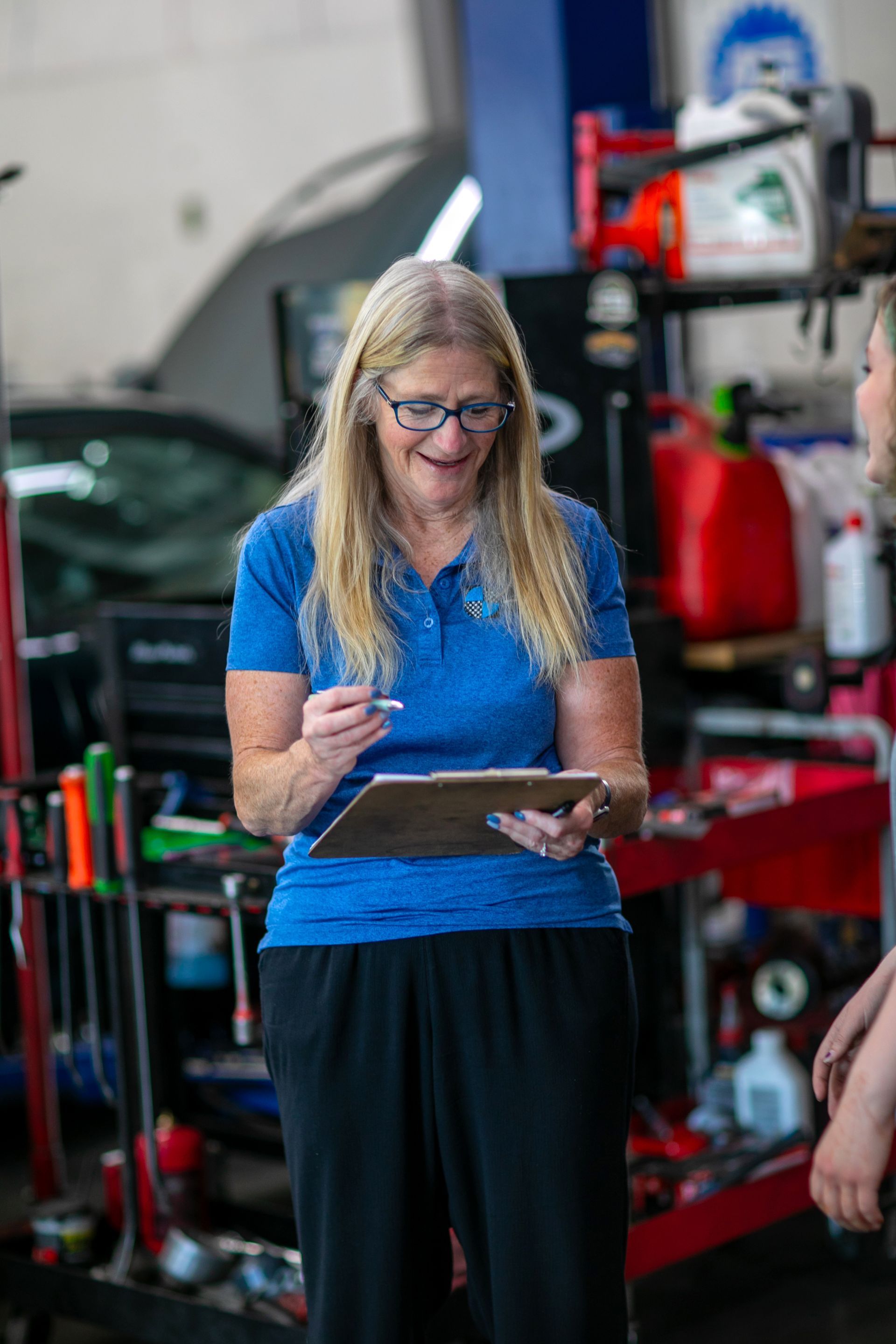 A person in a blue polo shirt and black pants smiles while holding a clipboard in an auto repair shop. | Bavarian Auto Haus