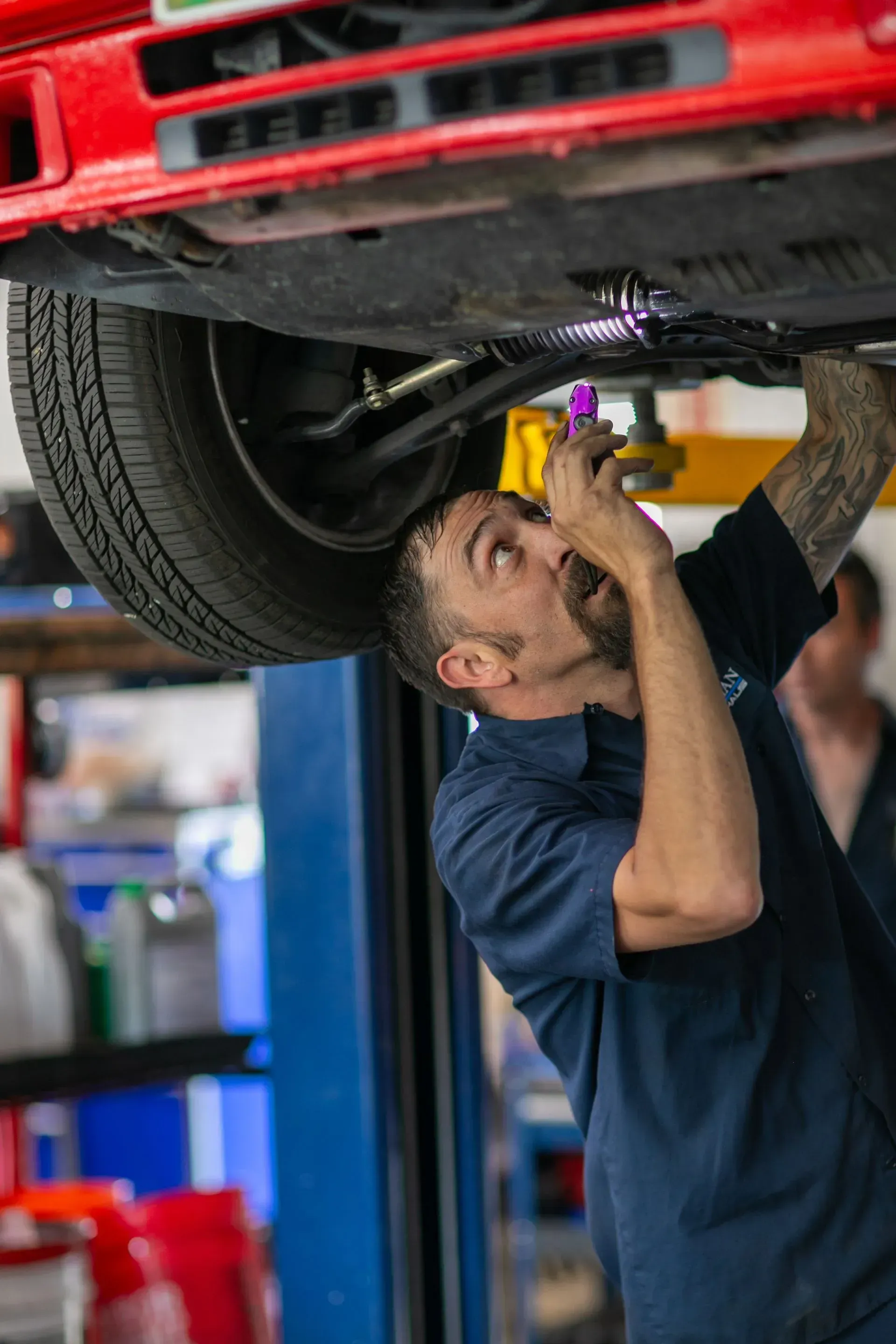 Mechanic working under a red car, using a flashlight. Workshop setting. | Bavarian Auto Haus