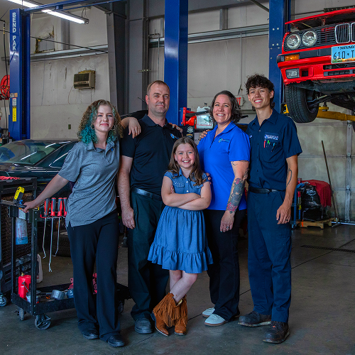 Family posing in auto repair shop; smiling, arms around each other, tools and car in background. | Bavarian Auto Haus