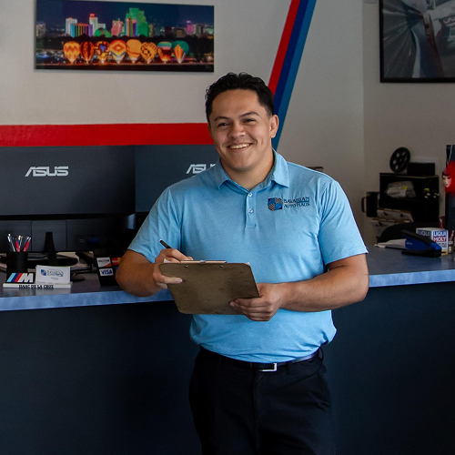 Man in blue polo, holding clipboard, smiles in auto shop. Red, white, and blue logo on shirt, desk behind him. | Bavarian Auto Haus