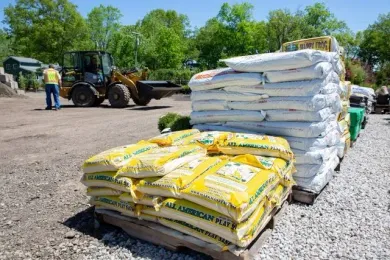 A worker stands near a front loader in a gravel lot next to stacked pallets of yellow and white landscape material bags.