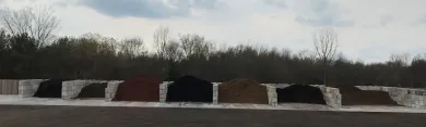 A row of various types and colors of mulch and soil stored in concrete bins outdoors against a tree line.