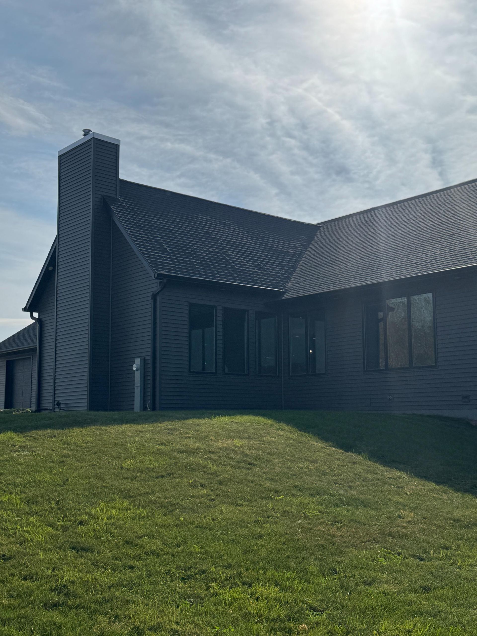 A dark-sided house with a prominent chimney, set against a bright, partly cloudy sky and a grassy lawn.