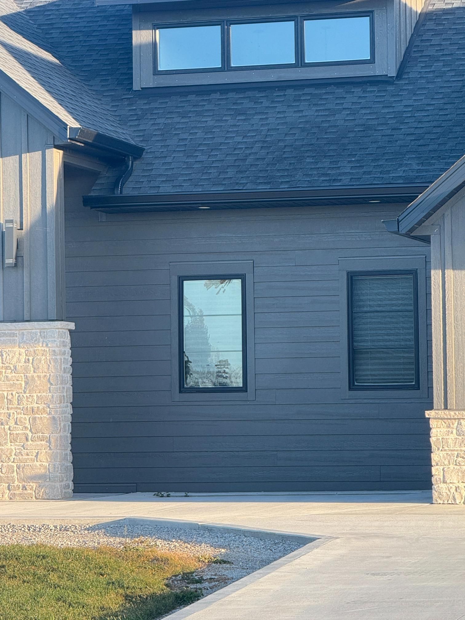 A modern house exterior with dark horizontal siding, stone pillars, a roof dormer, and two rectangular windows.