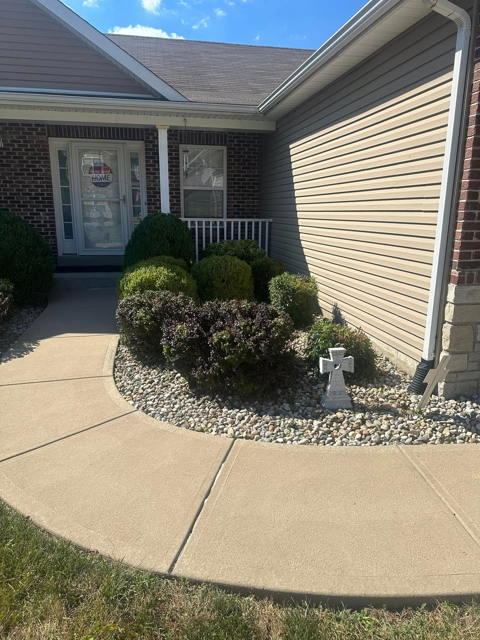 A curved concrete walkway leads to the front door of a brick and tan siding house, past a garden bed with shrubs and a cross.