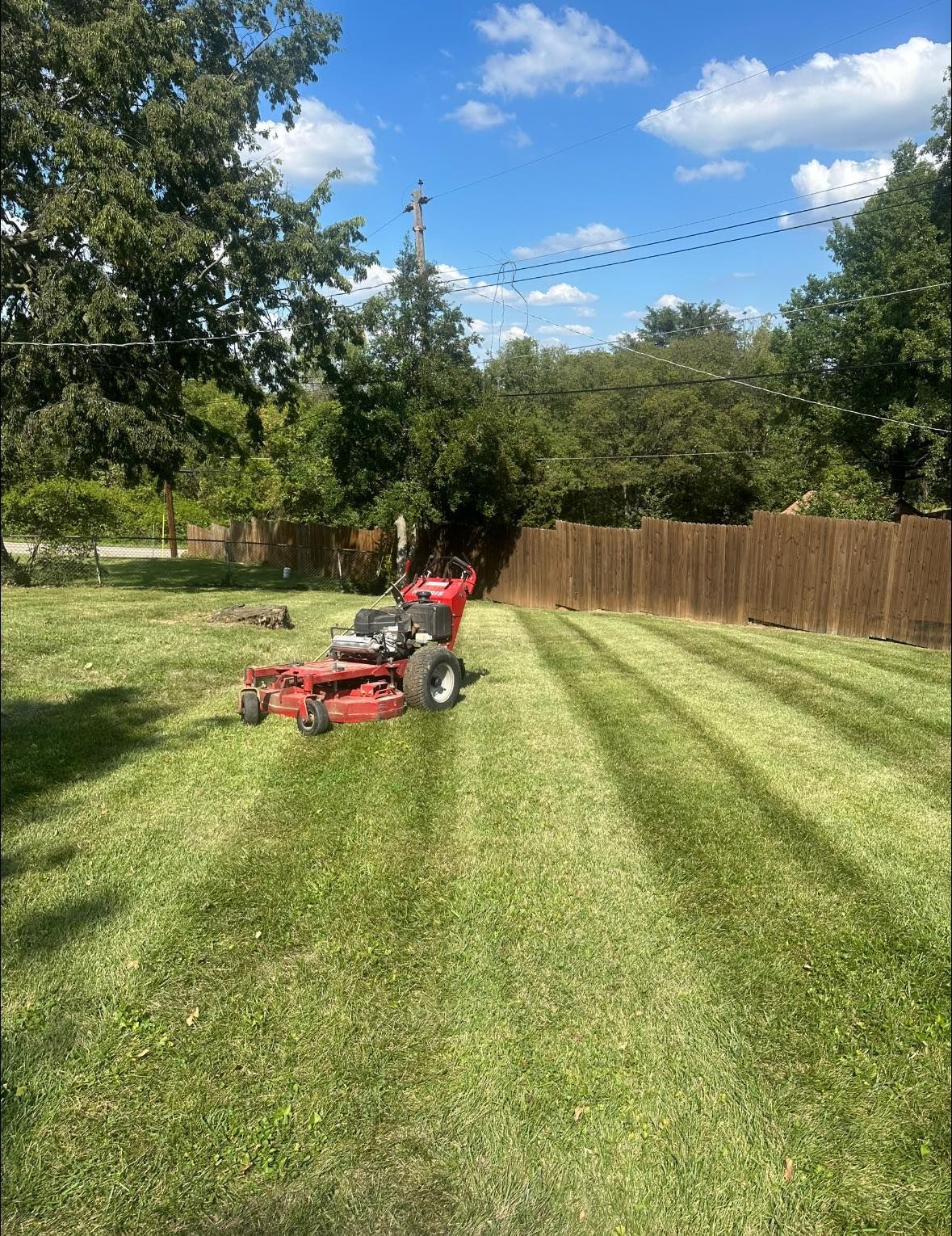 A red zero-turn mower cuts striped patterns into a green lawn in front of a wooden fence on a sunny day.
