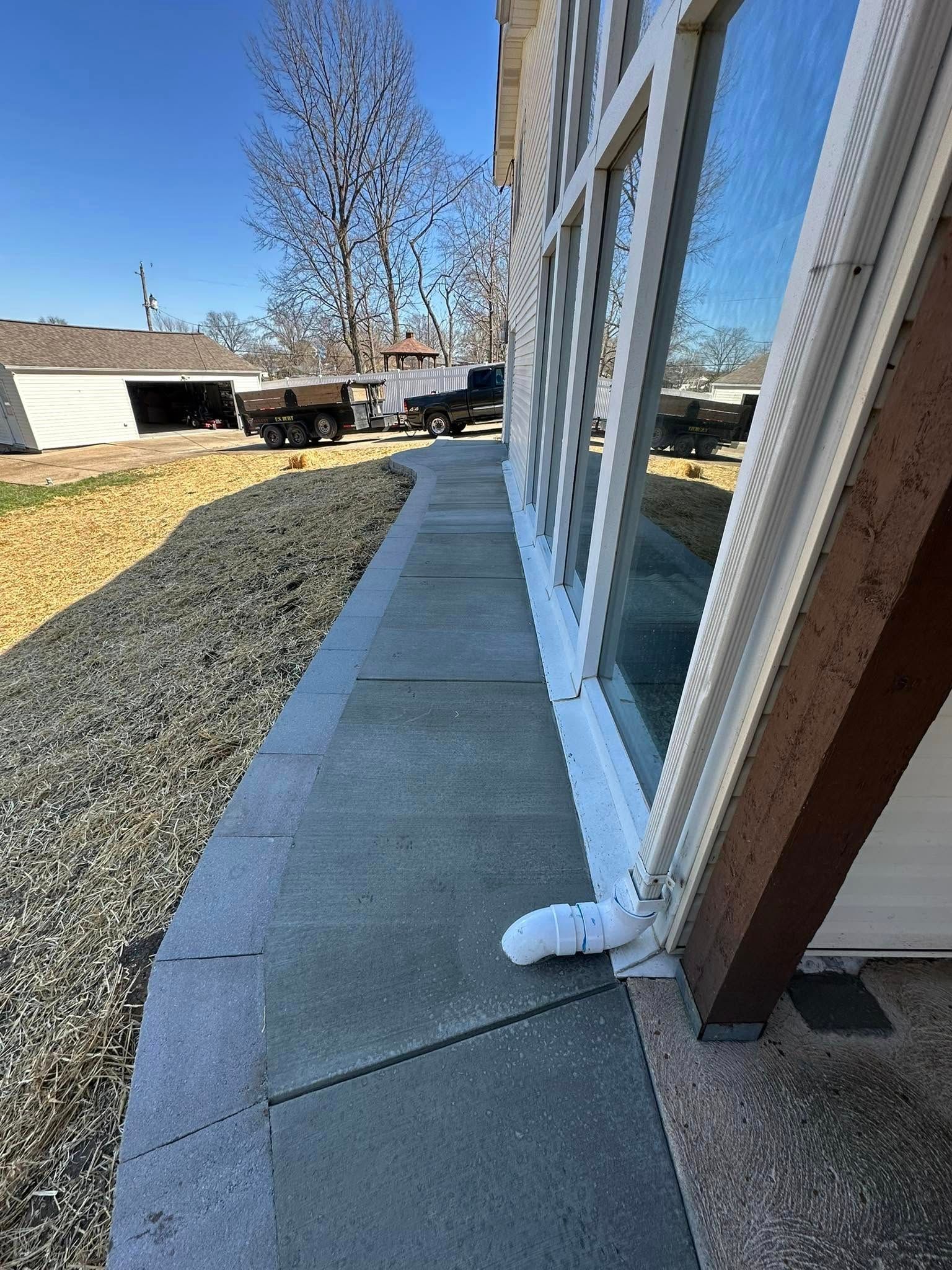A paved sidewalk runs alongside a building with large windows, next to a gravel area and a white outbuilding in the distance.