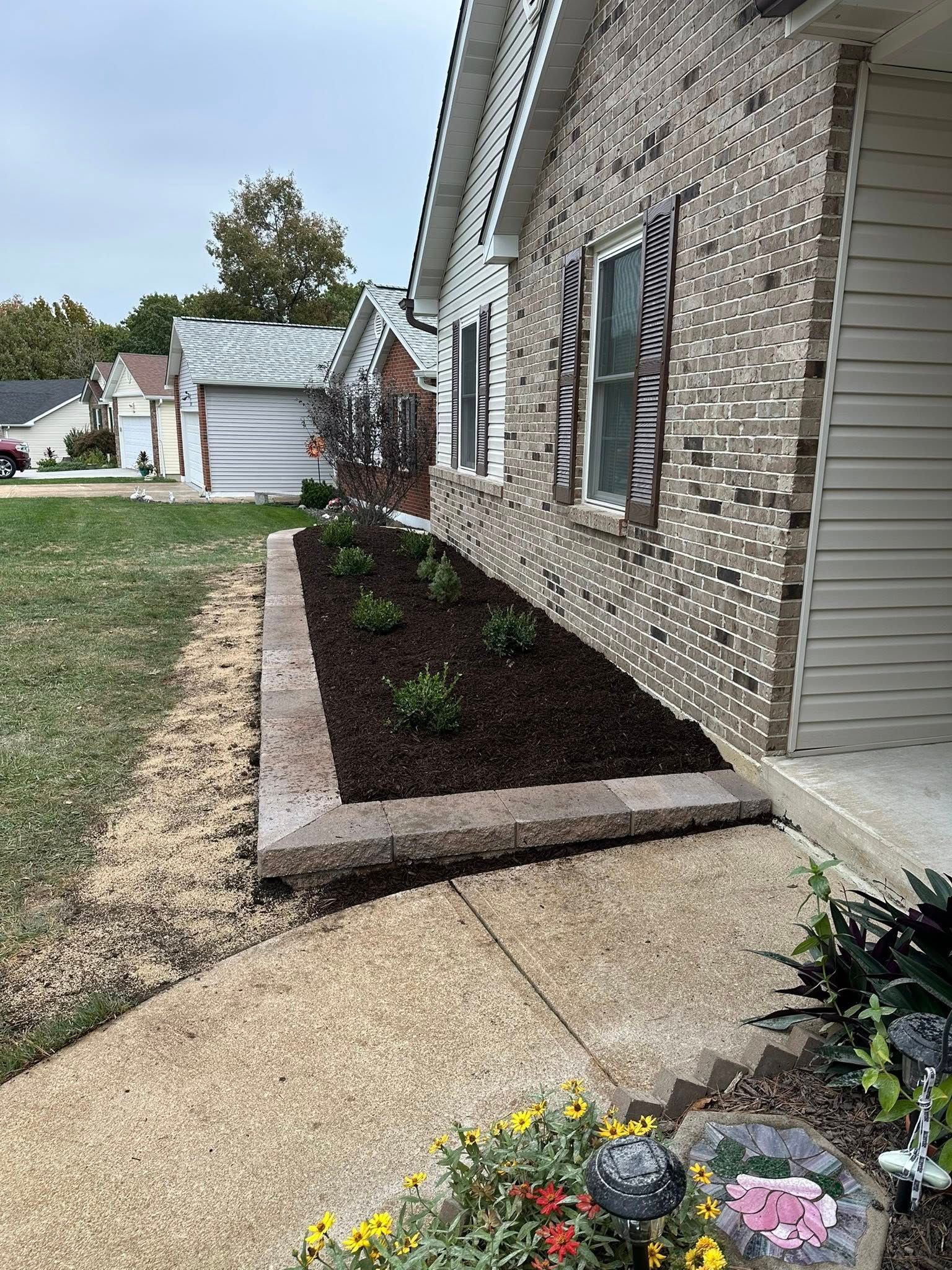 A brick house exterior featuring a newly landscaped rectangular garden bed bordered by concrete pavers with dark mulch.