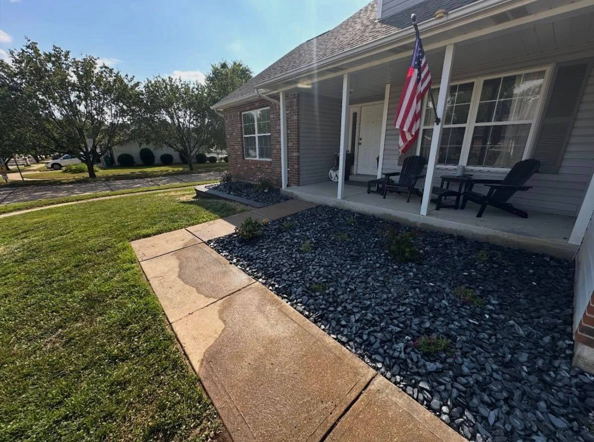 A front yard walkway leads to a house porch with dark rocks, two outdoor chairs, and an American flag.