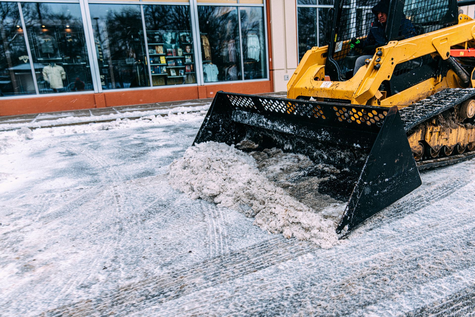 A yellow skid-steer loader plows a pile of slushy snow on a sidewalk in front of a storefront.