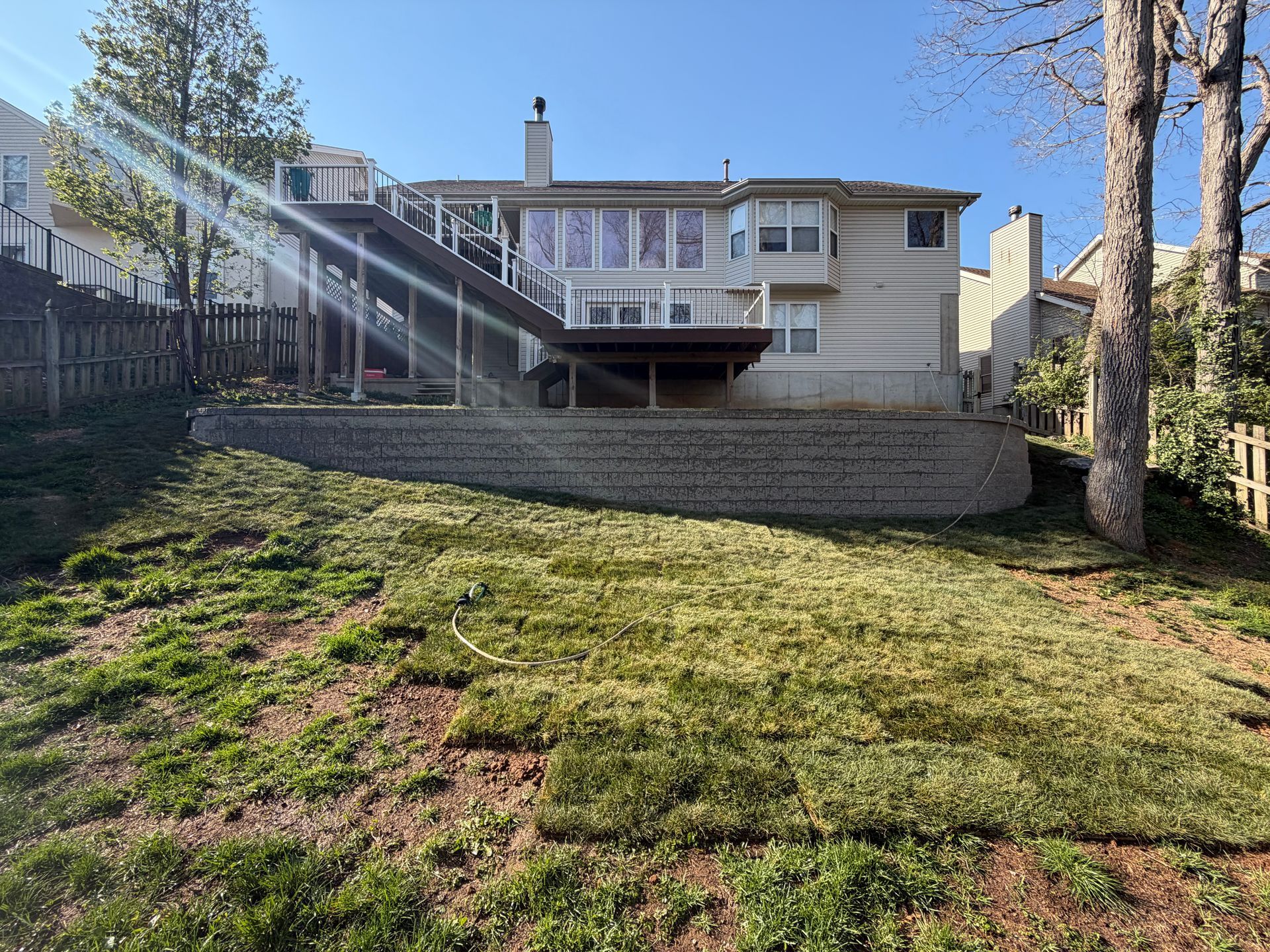 A beige two-story house with a large wooden deck stands above a stone retaining wall in a backyard with patchy grass.