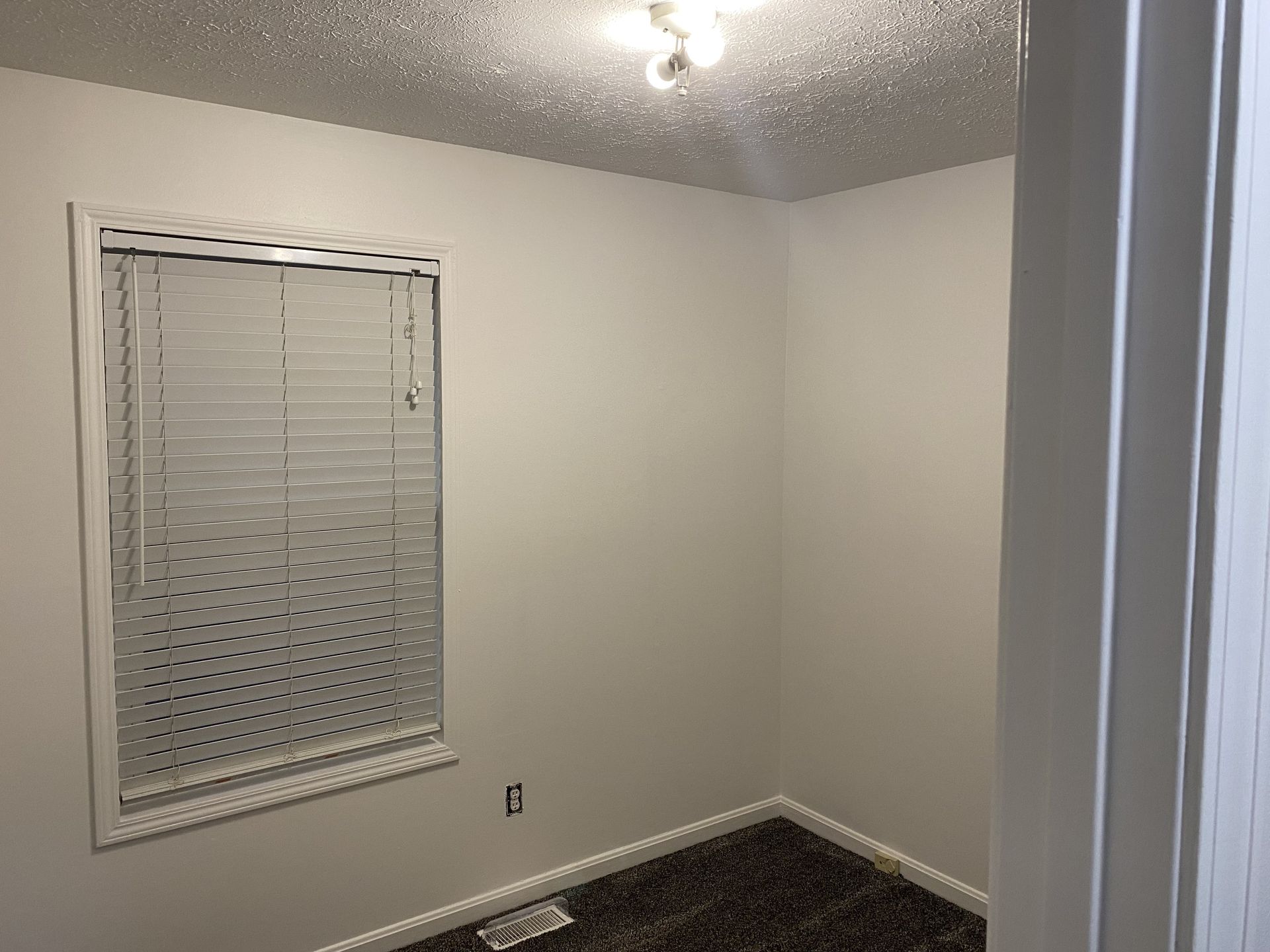 A small, empty bedroom with white walls, a single window with closed horizontal blinds, and dark carpet.