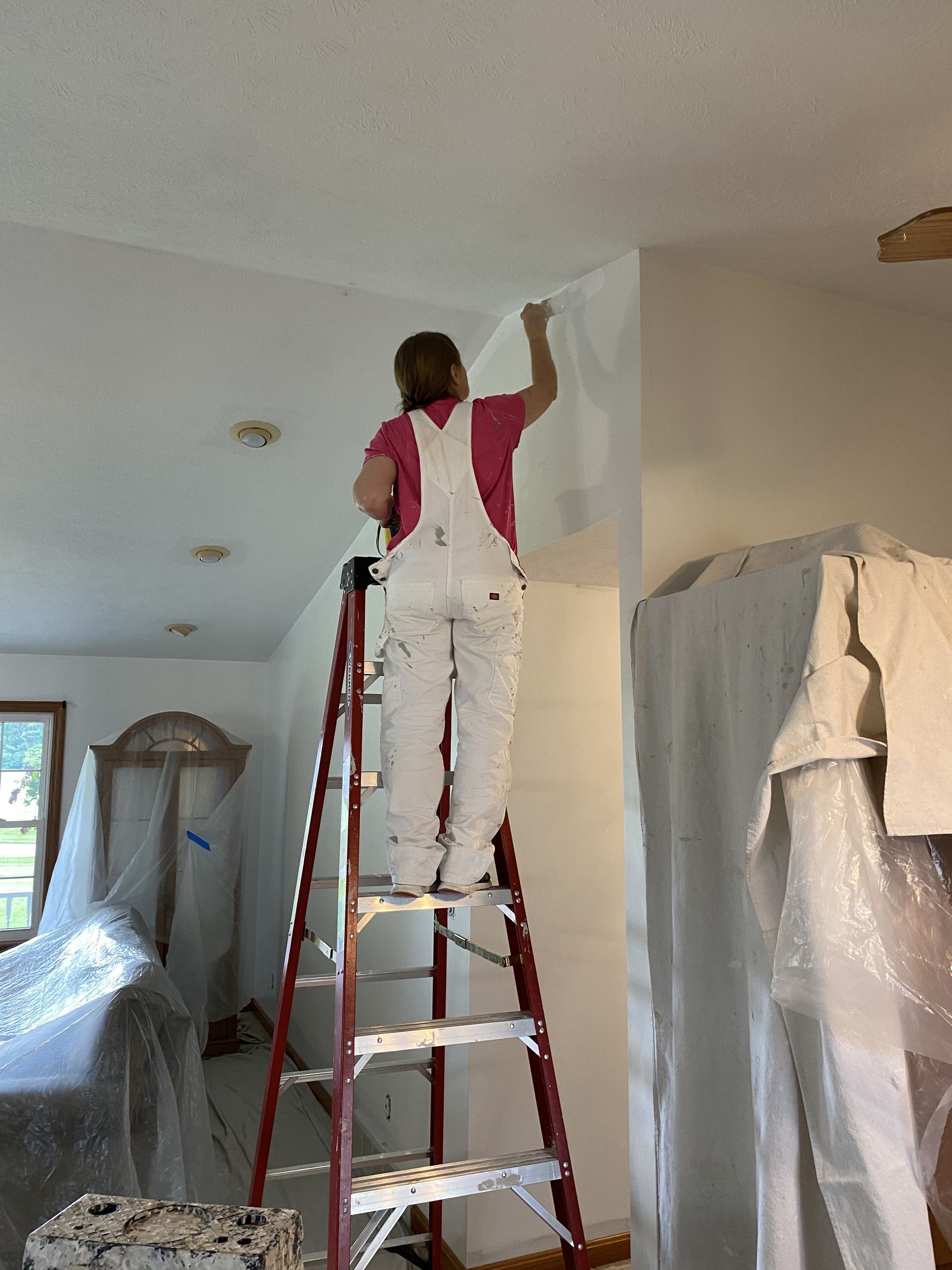 A person in white overalls standing on a red ladder painting the upper corner of a room wall white.