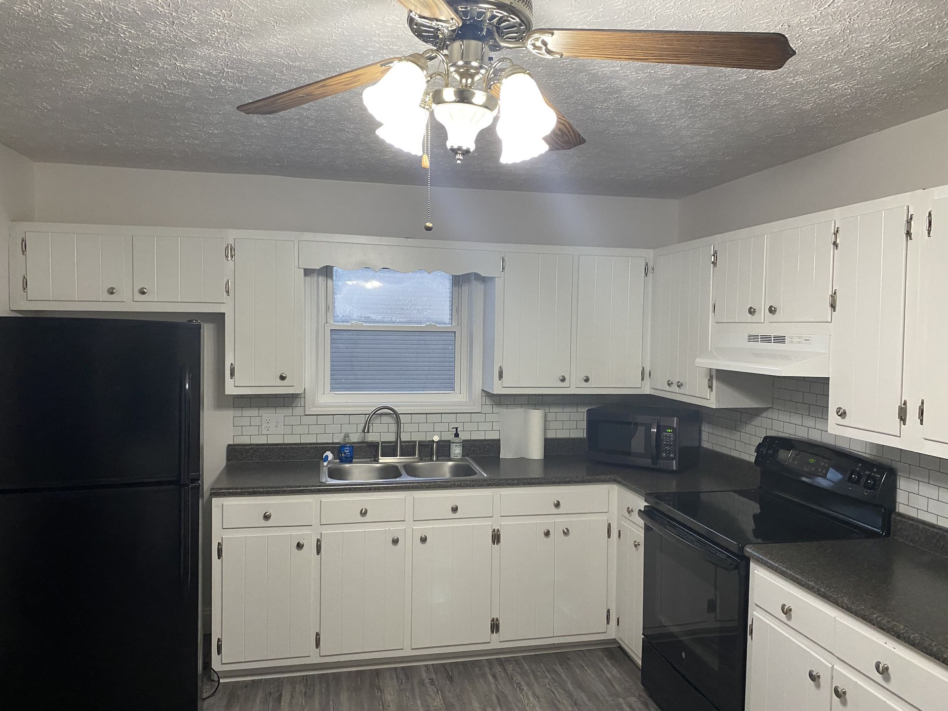 A kitchen with white cabinets, dark countertops, black appliances, a ceiling fan, and a window above the sink.