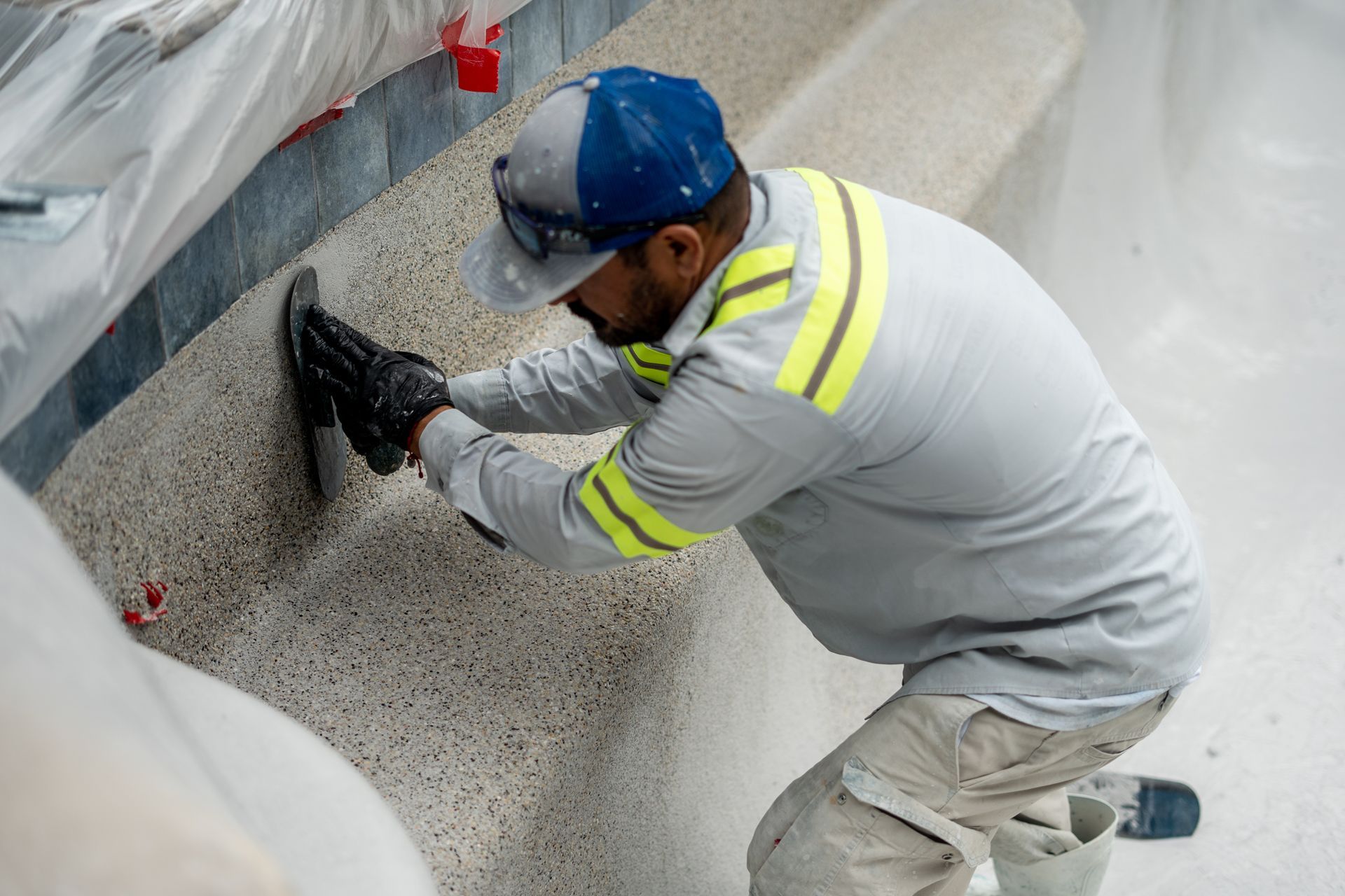 Man in work attire applying material to a pool wall, wearing a hat, gloves, and safety vest.