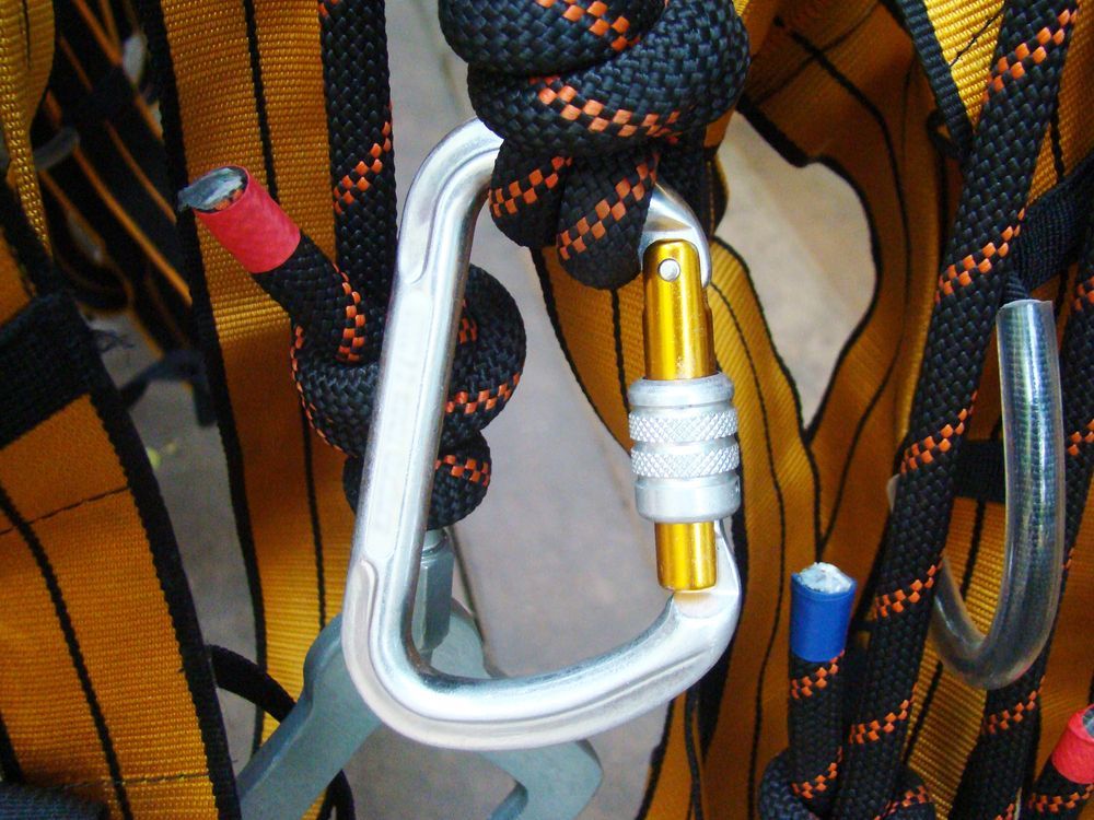 Primer plano de un mosquetón que conecta cuerdas negras y naranjas a un arnés amarillo.