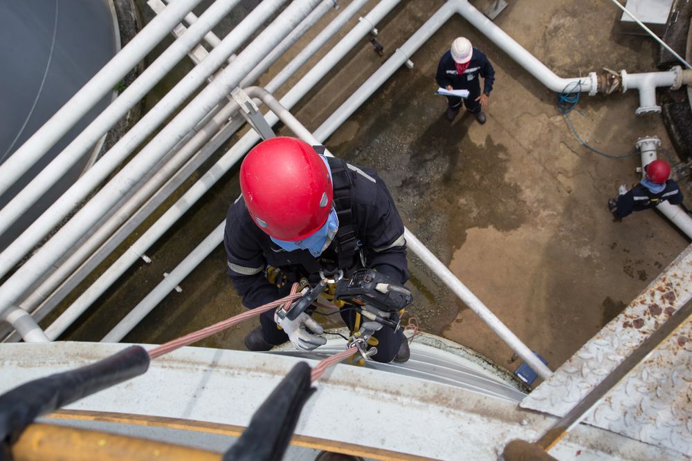 Un trabajador sujeto con arnés desciende en rappel por una estructura industrial; otros dos trabajadores observan.