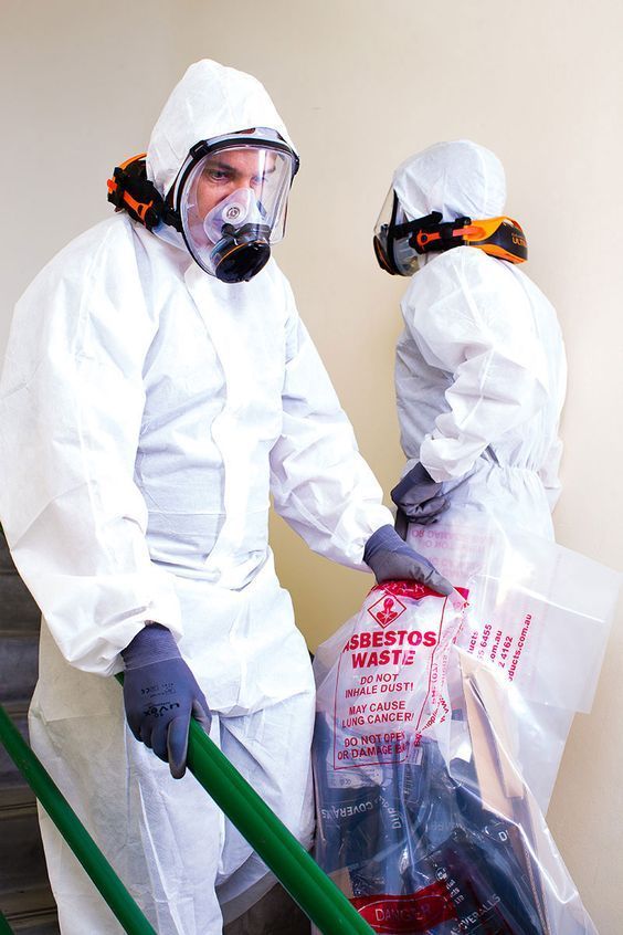 Dos trabajadores con trajes de protección química bajan escaleras abajo con bolsas de residuos de amianto.