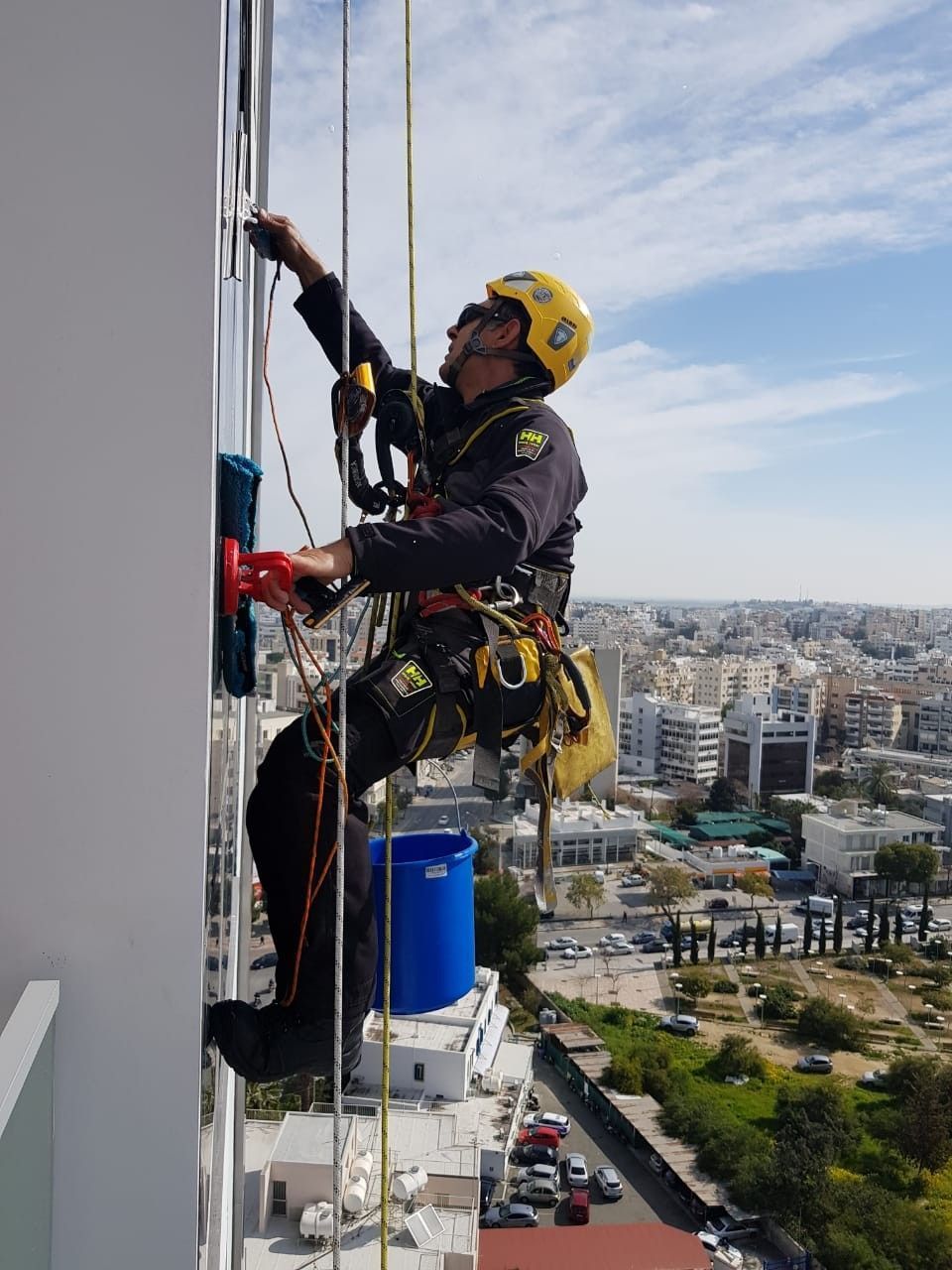 Limpiacristales en un rascacielos, limpiando cristales con una rasqueta, arnés, cubo azul, paisaje urbano de fondo.