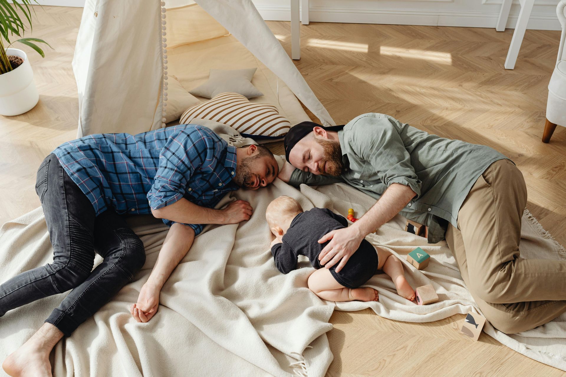 Two men and a baby are laying on a blanket on the floor.