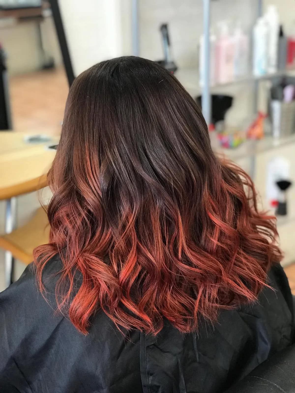Back view of medium-length wavy hair with a dark brown base transitioning into vibrant red ombre ends. — Devyani Hair & Beauty in Unanderra, NSW