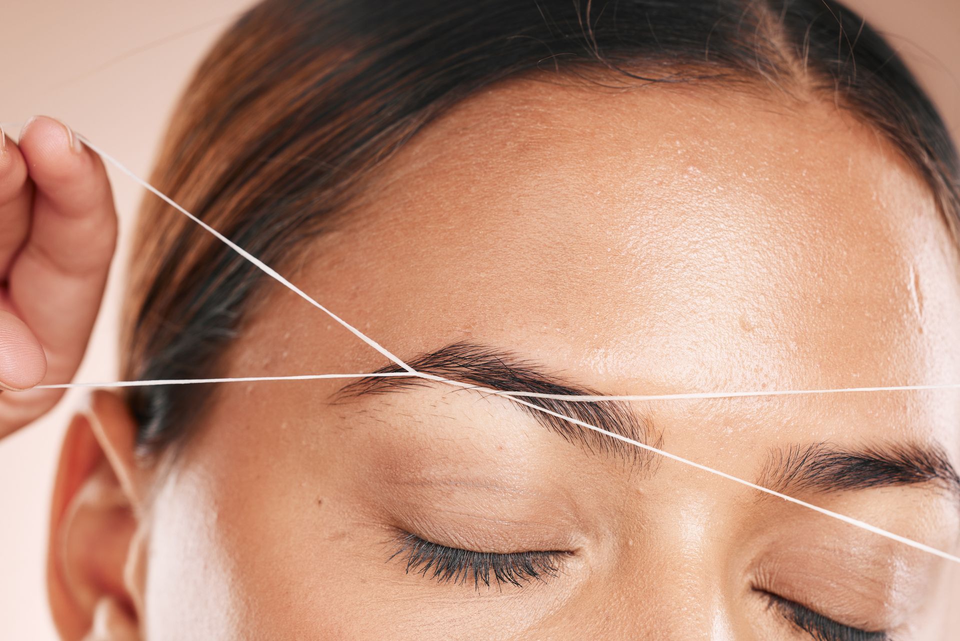 Eyebrow threading with white thread. Closed eyes, light brown skin, peach background.