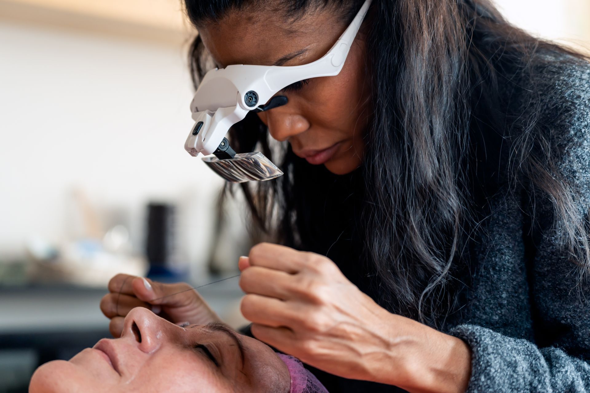Woman using magnifying glasses on a client's eyebrow, indoors.