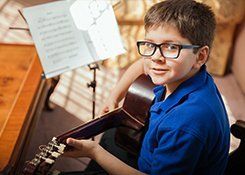 A little boy wearing glasses and a blue shirt, sitting holding a guitar behind a music stand