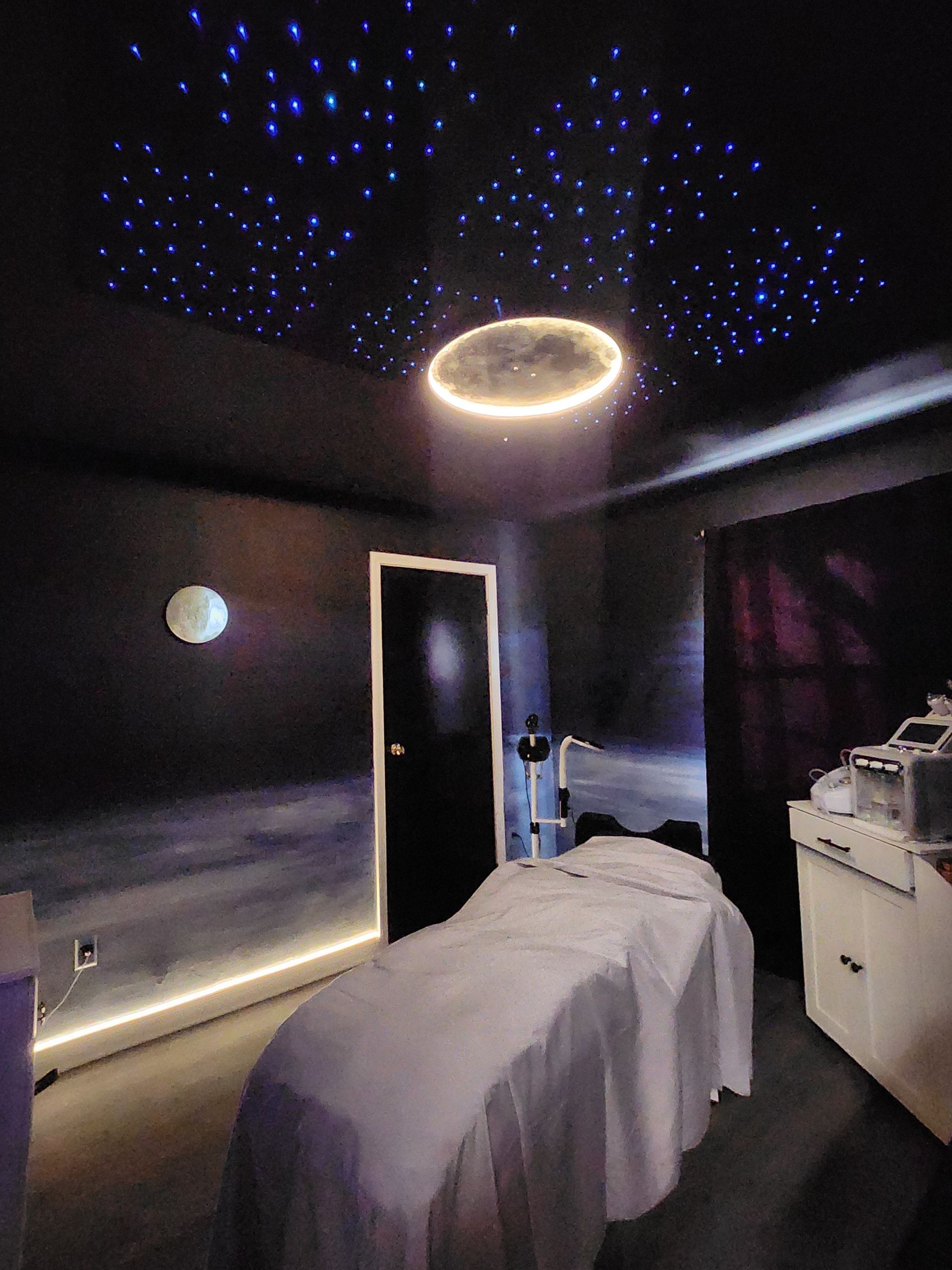 Relaxation room with a starry sky ceiling, glowing ring, and treatment bed. Dark walls with subtle lighting and equipment.