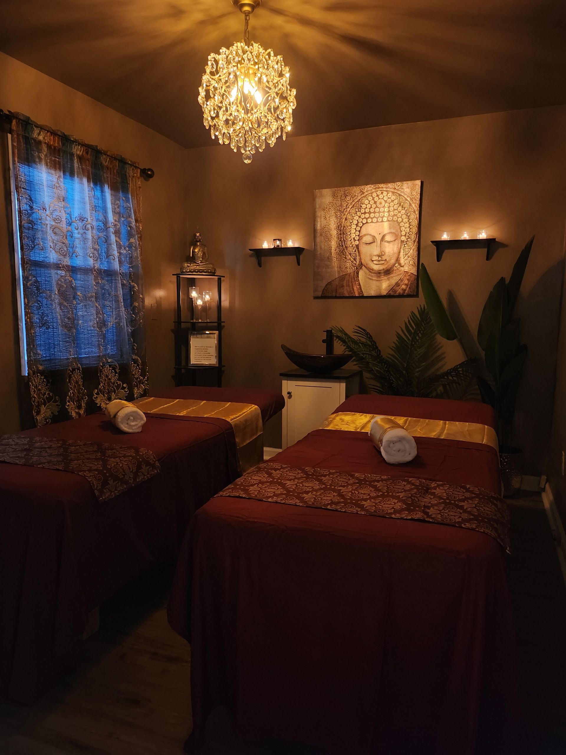 Spa room with two massage tables covered in red, lit by candles and a chandelier. Buddha art on wall.