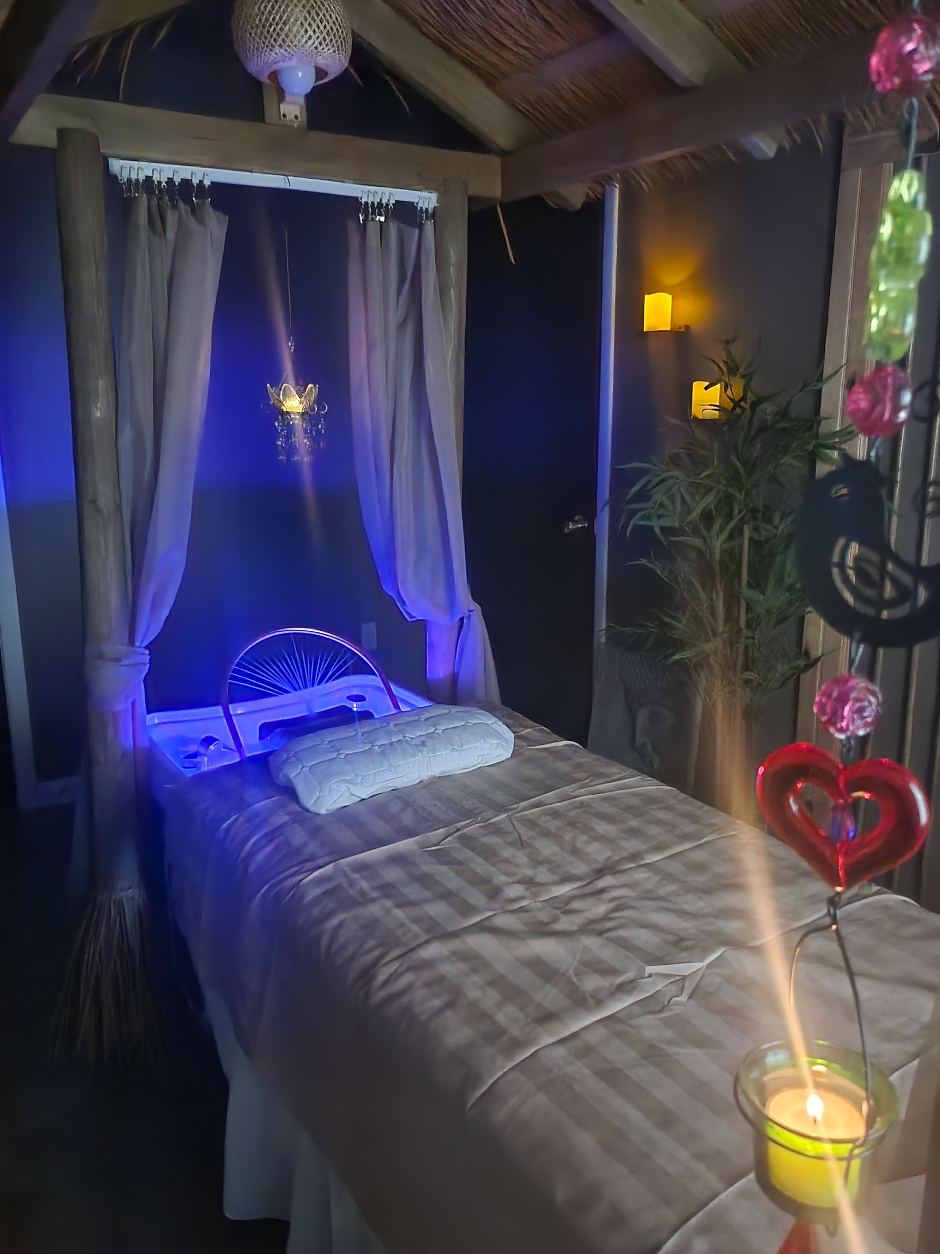 Spa treatment room with bed, drapes, and soft lighting; decorated with bamboo, candles, and heart-shaped decor.