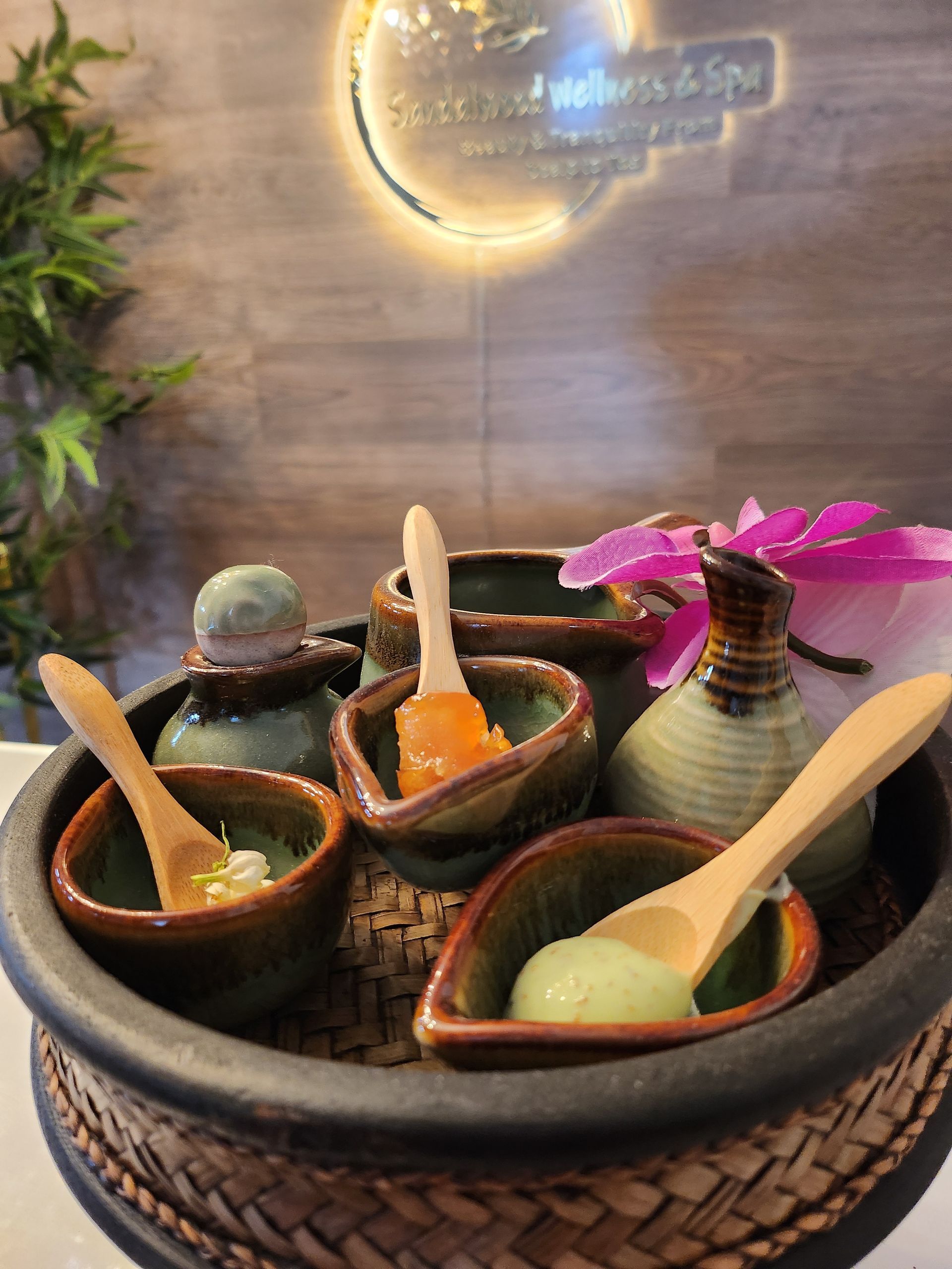 Spa treatment set: small ceramic bowls with colorful scrubs, wooden spoons, and a hot compress on a tray.