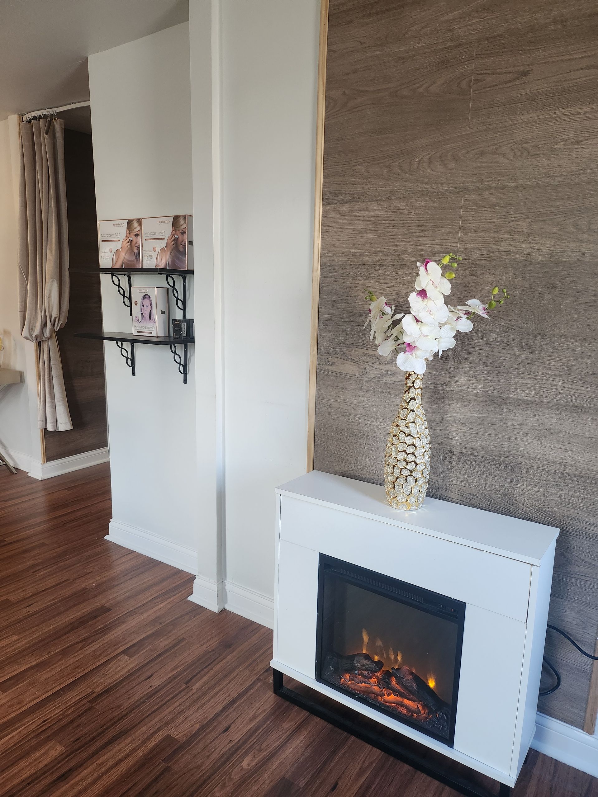A white electric fireplace with a vase of flowers on top, set against a textured stone wall and hardwood floors.