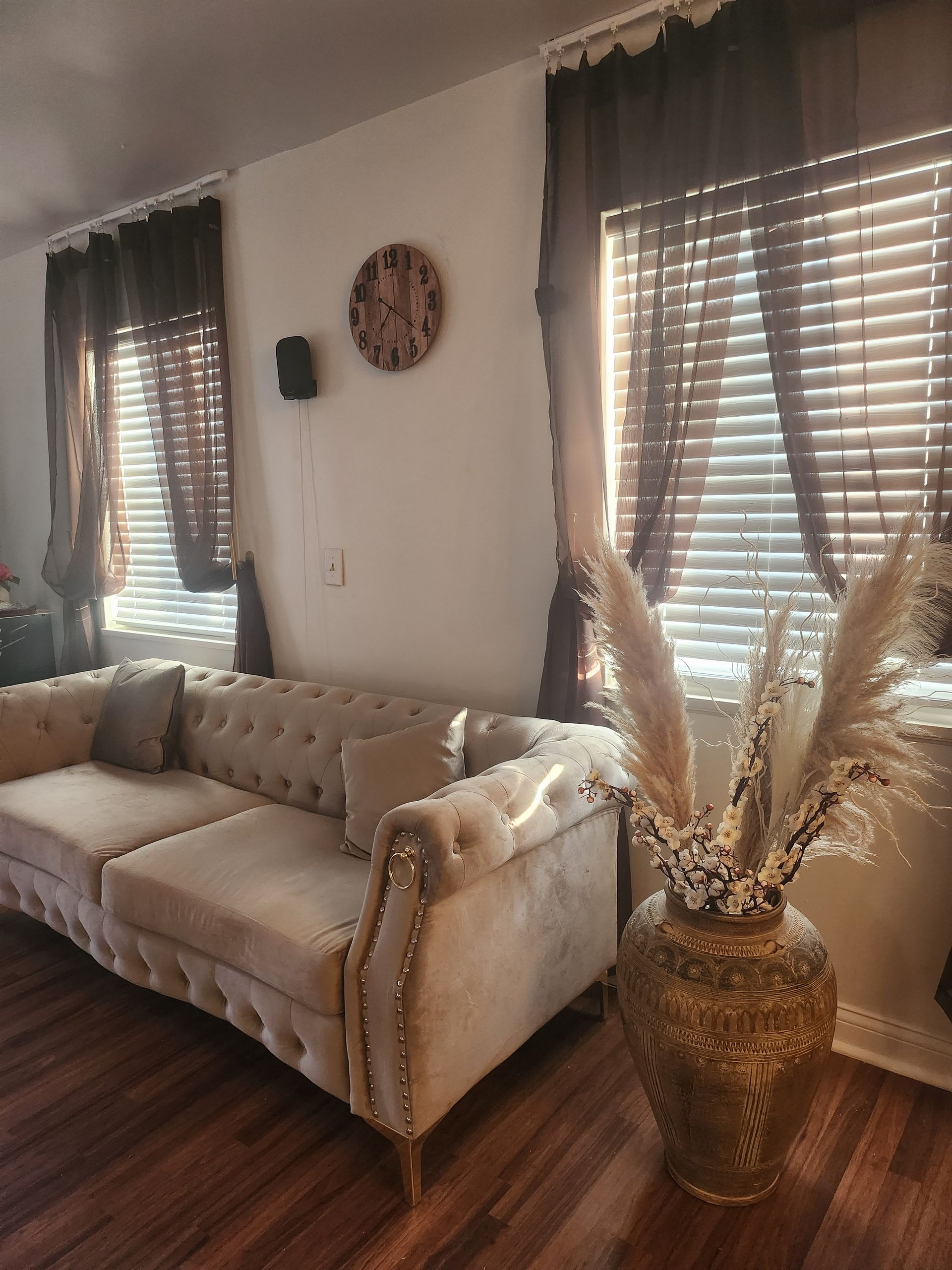 Beige tufted sofa in a room with wood floors, dark brown curtains, a decorative clock, and a gold vase with pampas grass.