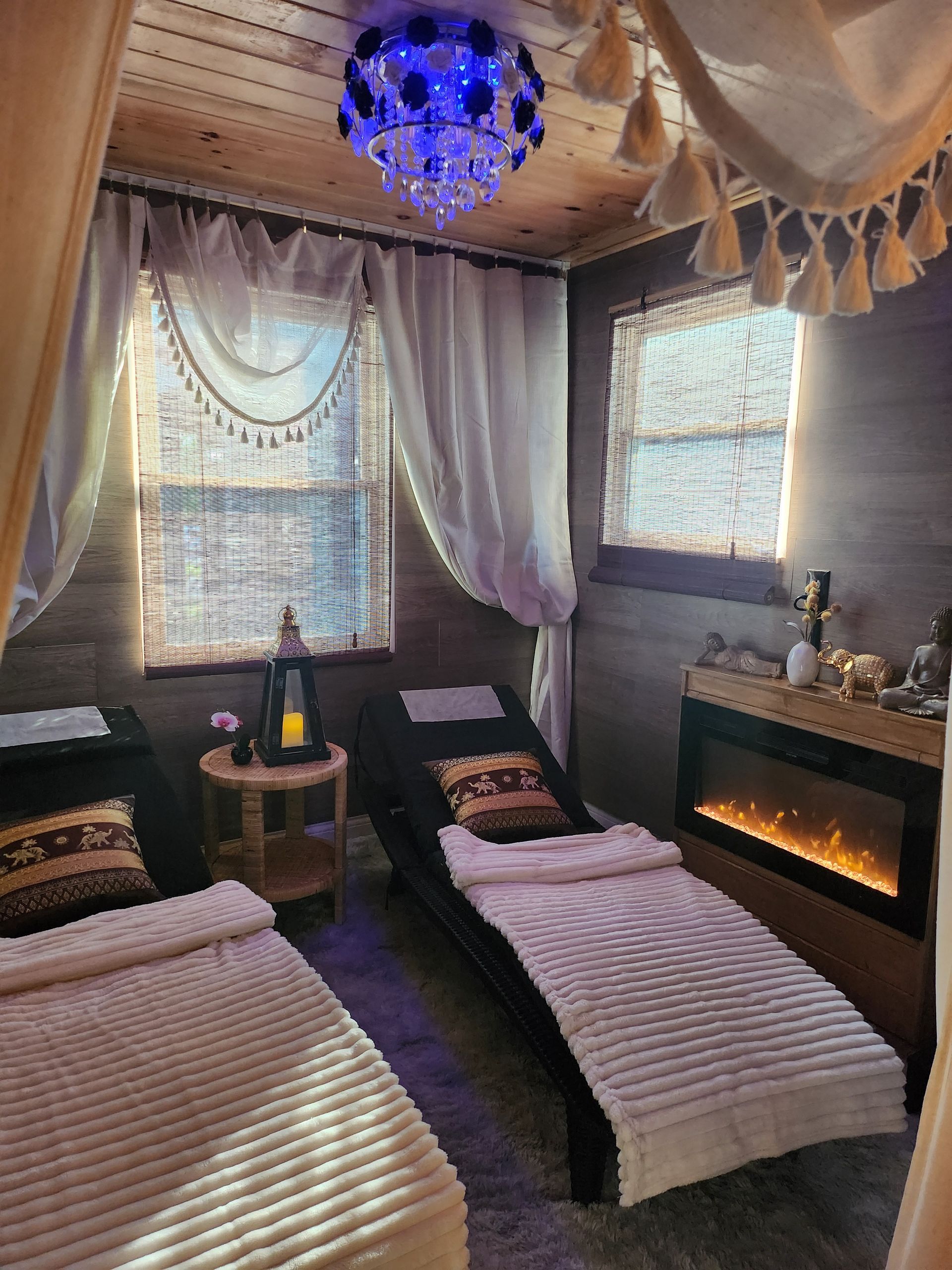 A serene spa room with two massage beds, curtains, and a fireplace. A blue chandelier hangs overhead.