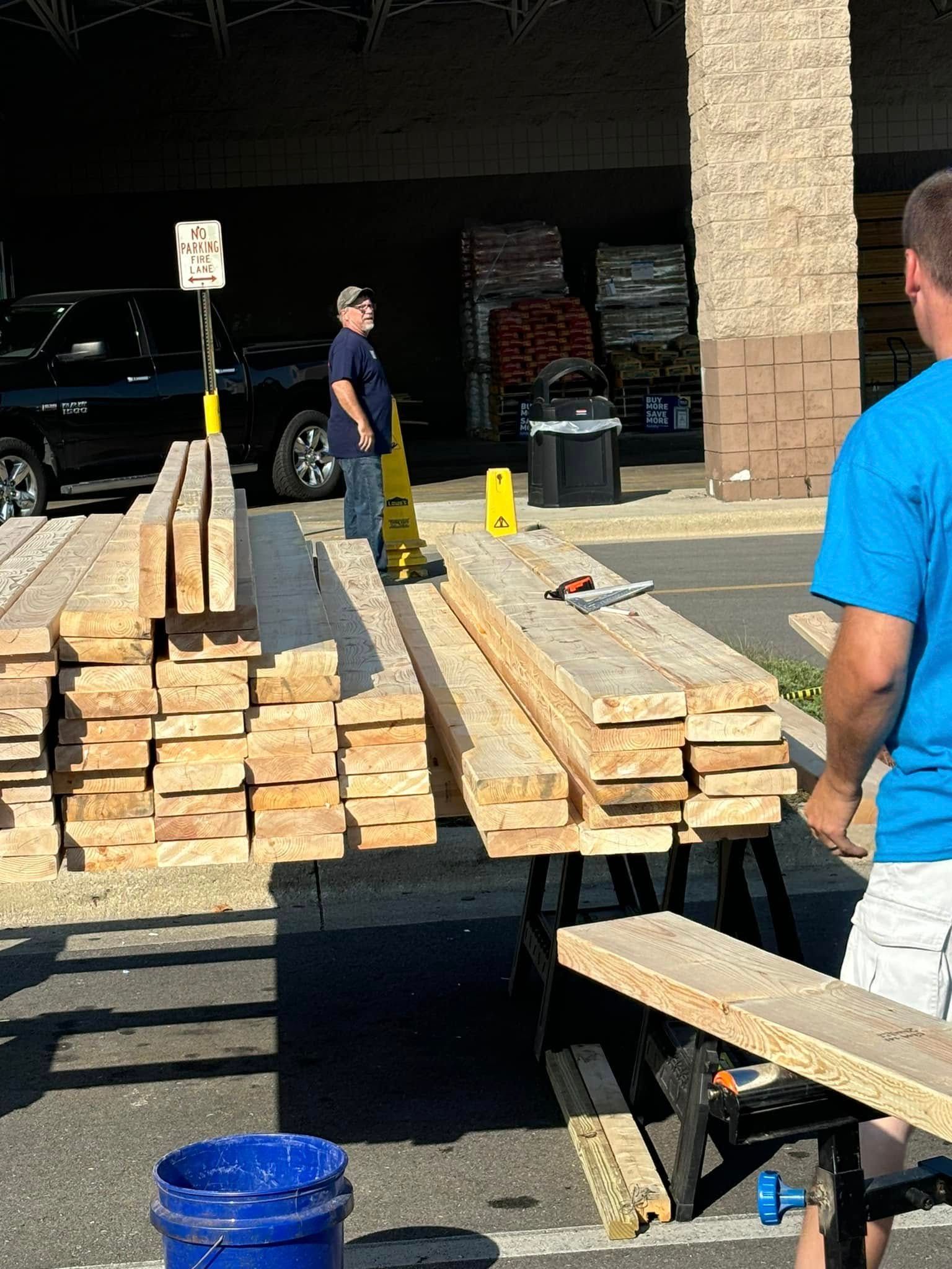 A man in a blue shirt is standing next to a pile of wood.