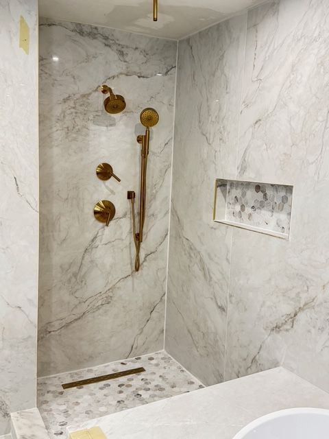 Shower with marble-look tile, gold fixtures, built-in shelf, and pebble-tiled floor.