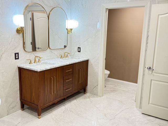 Bathroom with a wooden vanity, marble-look walls, gold fixtures, and two arched mirrors.