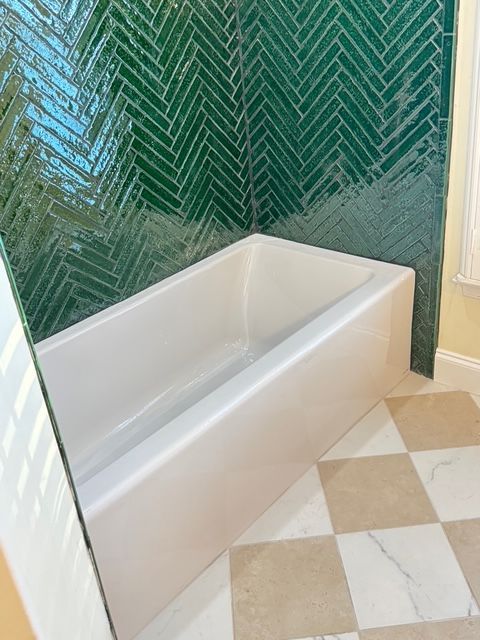 White bathtub against a green herringbone-tiled wall in a bathroom with checkered beige and white floor tiles.